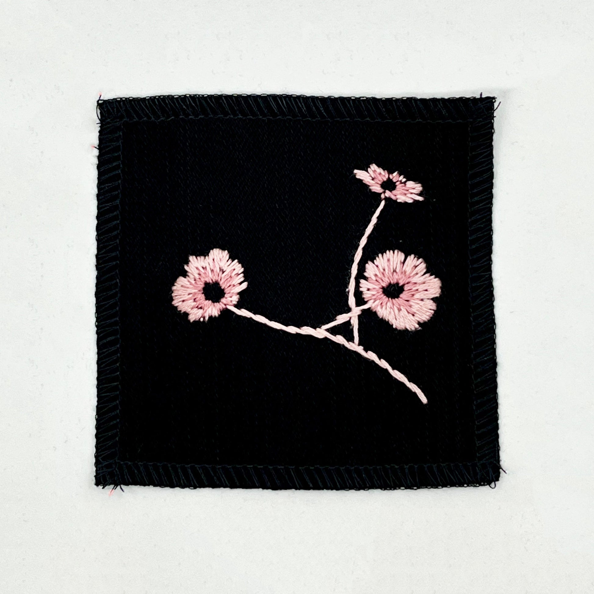 a black colored square patch, hand embroidered with three poppies on a stem, in shades of pink, with overlocked edges, on a white background