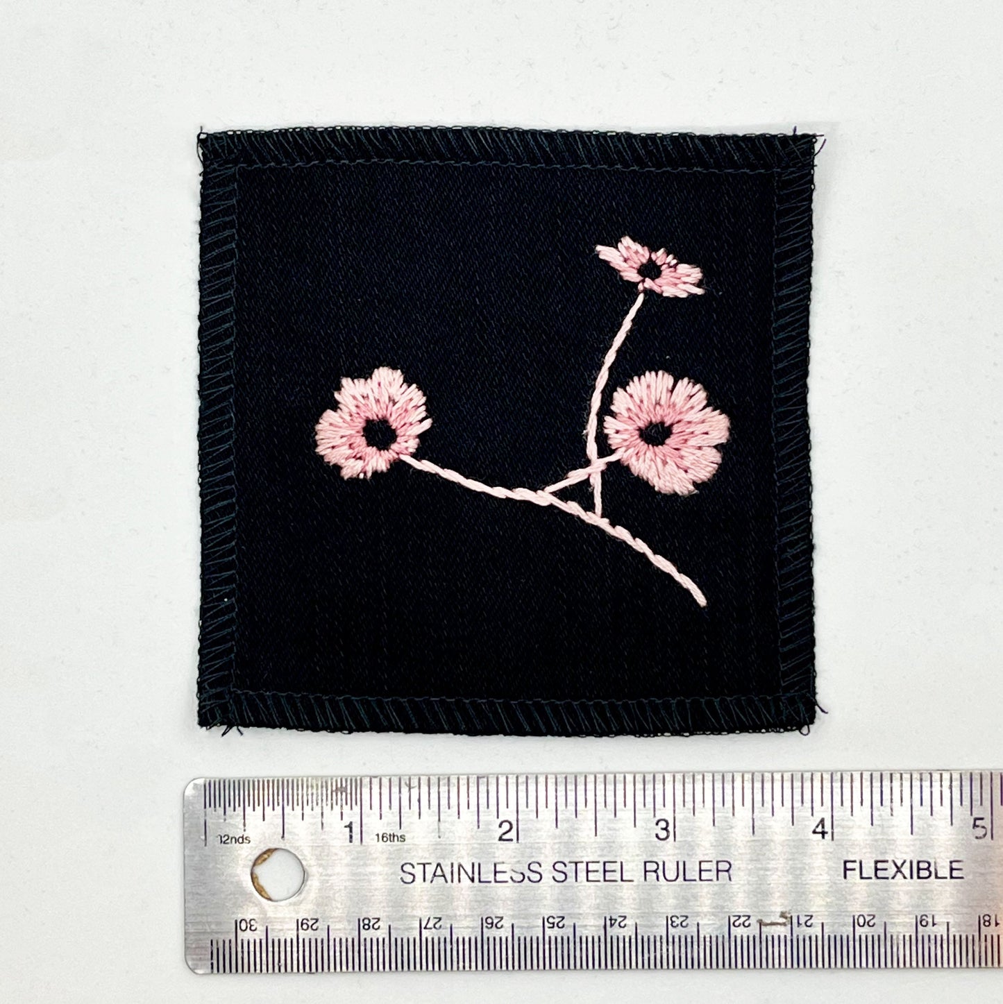 a black square patch, hand embroidered with three poppies on a stem, in shades of pink, with overlocked edges, placed next to a metal ruler to show a width of four inches, on a white background