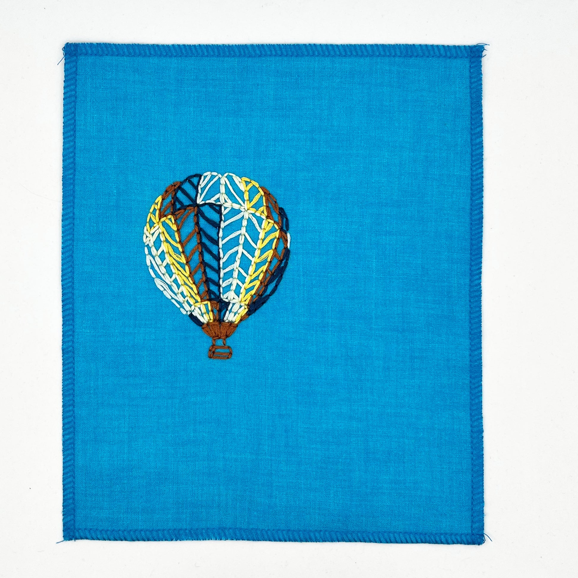 a bright blue fabric wall hanging with a hot air balloon stitched on it off center, in dark blue, light blue, light green, yellow and rust colored thread, on a white background