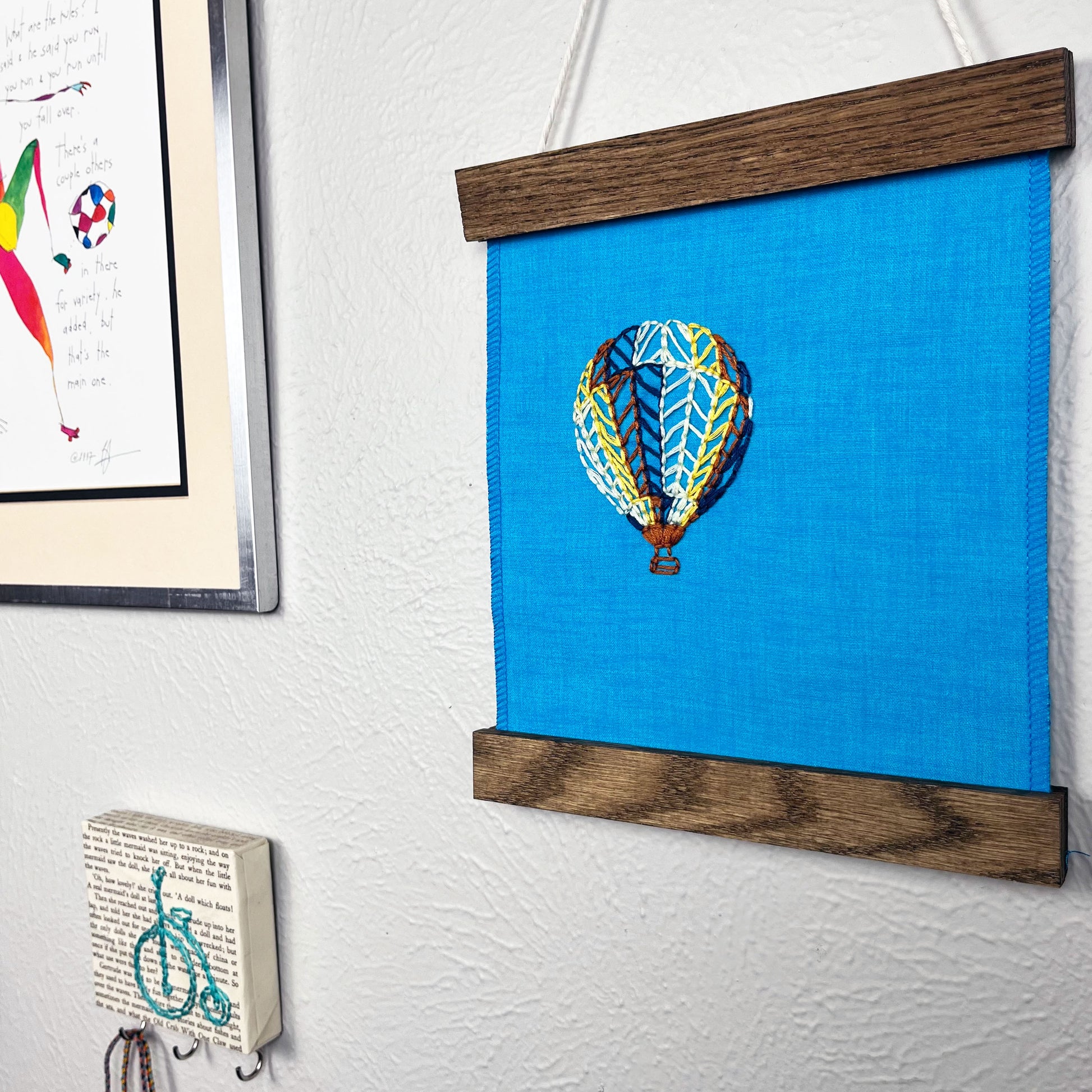 a bright blue fabric wall hanging in a magnetic wood frame, with a hot air balloon stitched on it off center, in dark blue, light blue, light green, yellow and rust colored thread, hanging on a wall near other colorful artwork