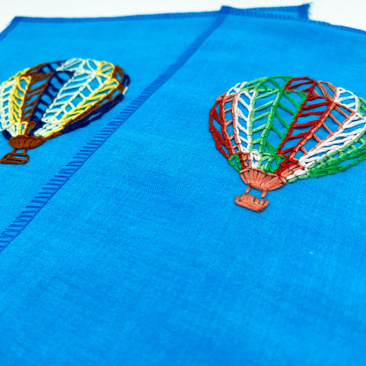 Close up angled view of two bright blue fabric wall hangings with hot air balloons stitched on them, one in dark colors one in bright colors