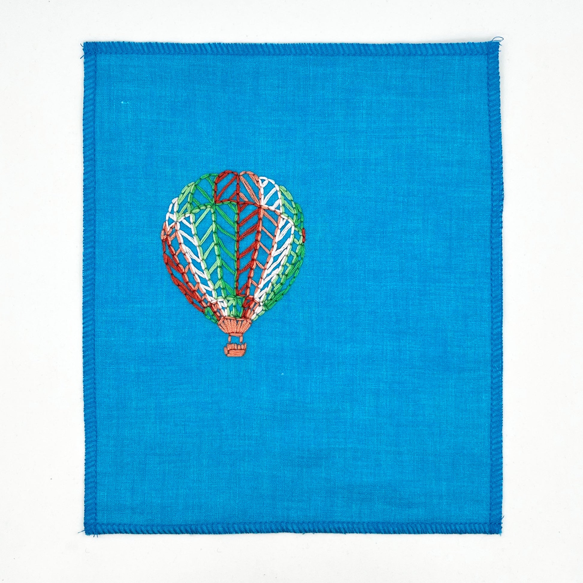 a bright blue fabric wall hanging with a hot air balloon stitched on it off center, in red, pink, light pink, light green and dark green colored thread, on a white background