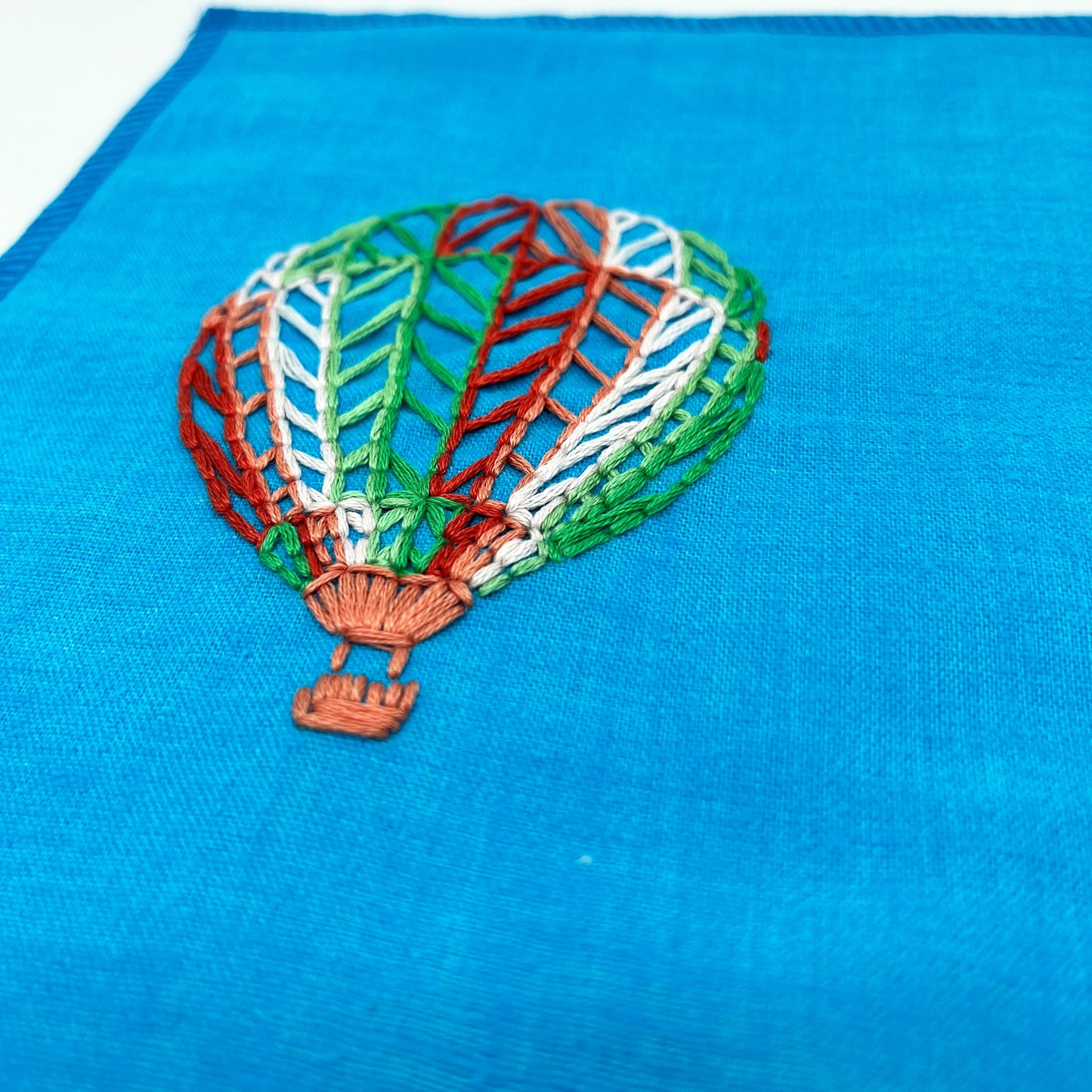 a close up angled view of a bright blue fabric wall hanging with a hot air balloon stitched on it off center, in red, pink, light pink, light green and dark green colored thread, on a white background