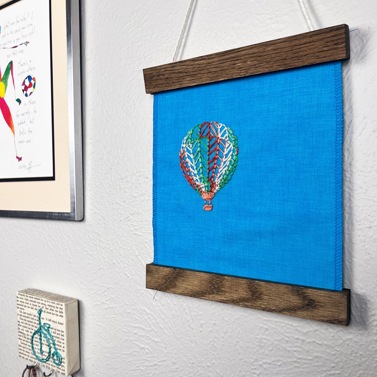 a bright blue fabric wall hanging in a magnetic wood frame, with a hot air balloon stitched on it off center, in red, pink, light pink, light green and dark green colored thread, hanging on a wall near other colorful artwork