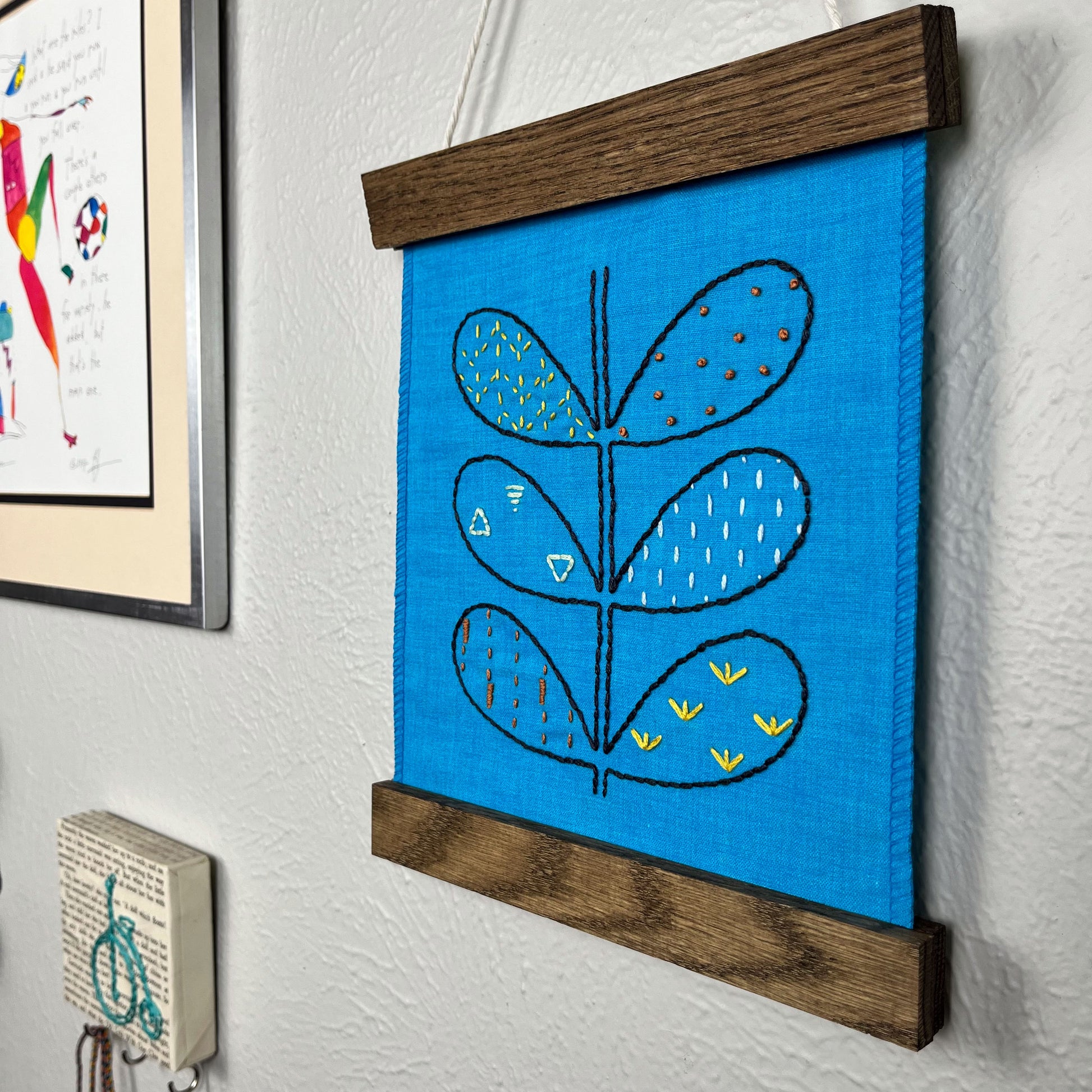 a small bright blue fabric wall hangings, in a magnetic wood frame, hand embroidered with the outline of symmetrical petals on a stem, each petal filled with different stitches like french knots, running stitches and triangles, in different colors