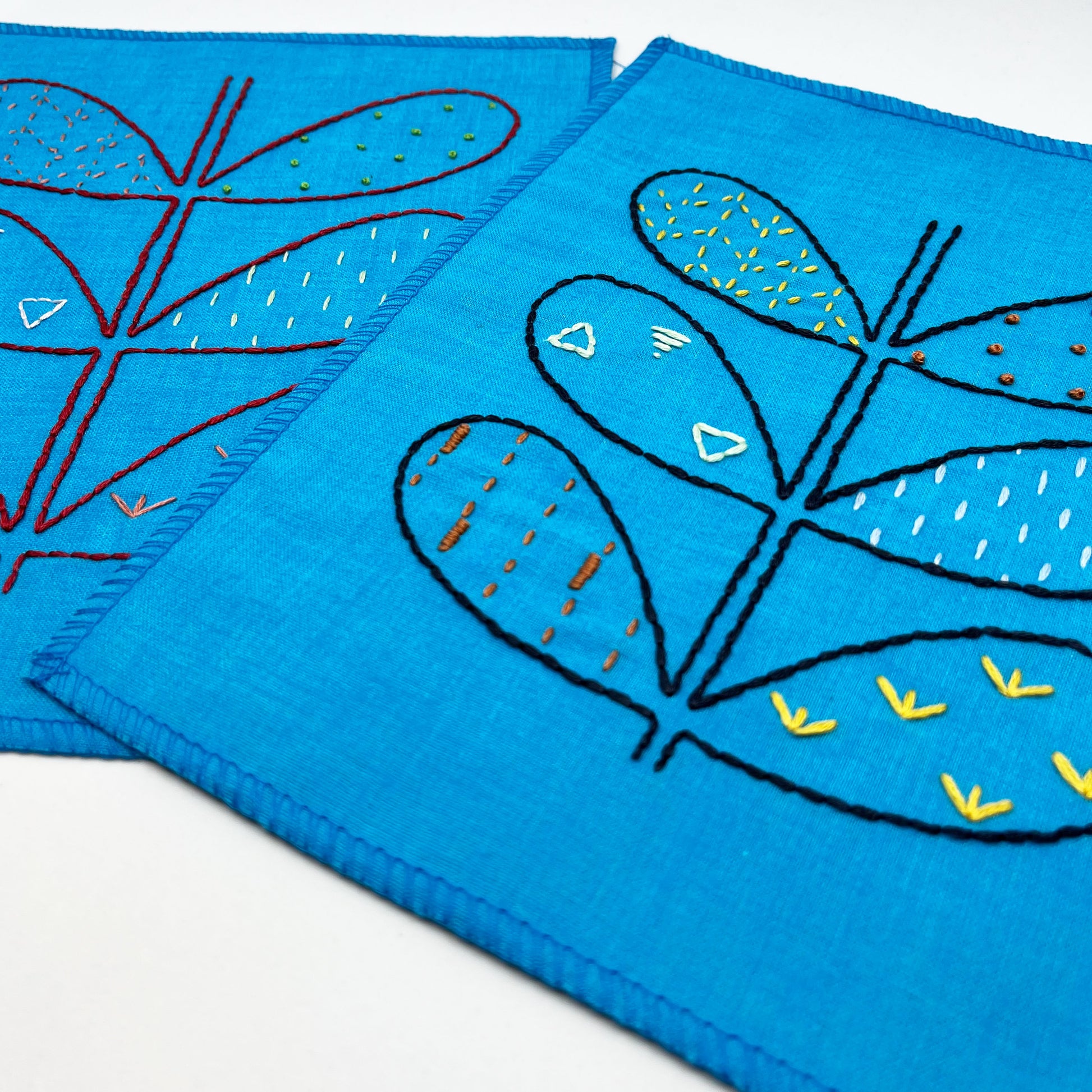 two small bright blue fabric wall hangings, hand embroidered with the outline of symmetrical petals on a stem, each petal filled with different stitches like french knots, running stitches and triangles, in different colors, on a white background