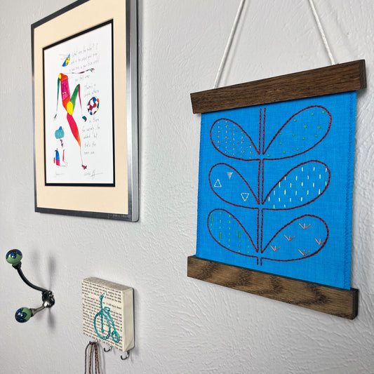 a small bright blue fabric wall hangings, in a magnetic wood frame, hand embroidered with the outline of symmetrical petals on a stem, each petal filled with different stitches like french knots, running stitches and triangles, in different colors