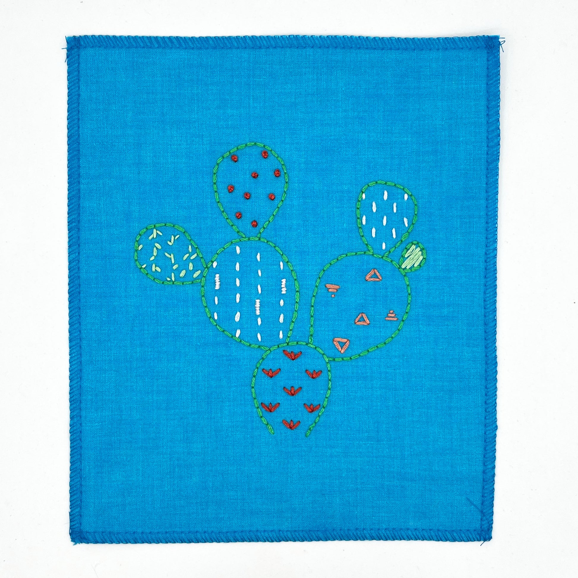 a small bright blue fabric wall hanging, hand embroidered with the outline of a prickly pear cactus in green, each pad filled with different stitches like french knots, running stitches and triangles, in different colors, on a white background