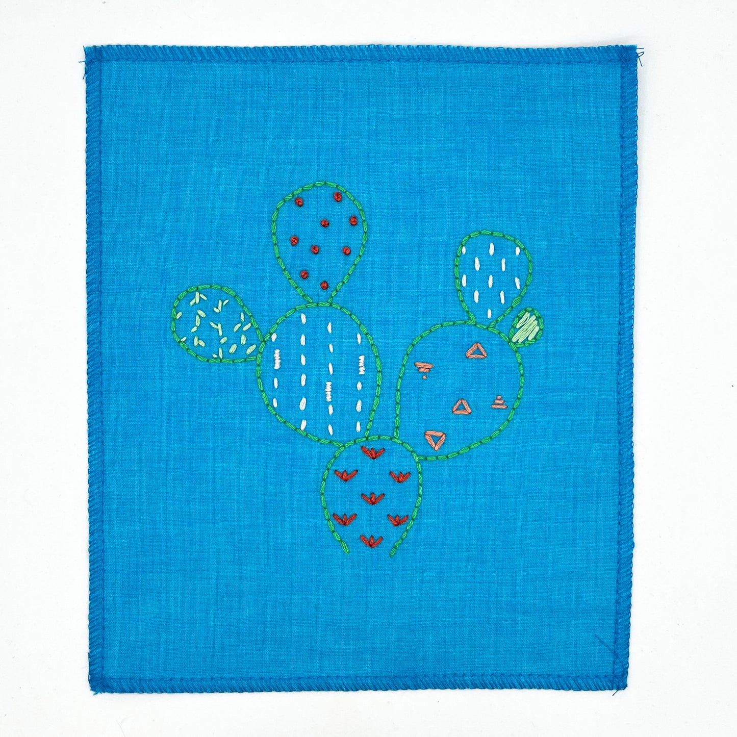 a small bright blue fabric wall hanging, hand embroidered with the outline of a prickly pear cactus in green, each pad filled with different stitches like french knots, running stitches and triangles, in different colors, on a white background