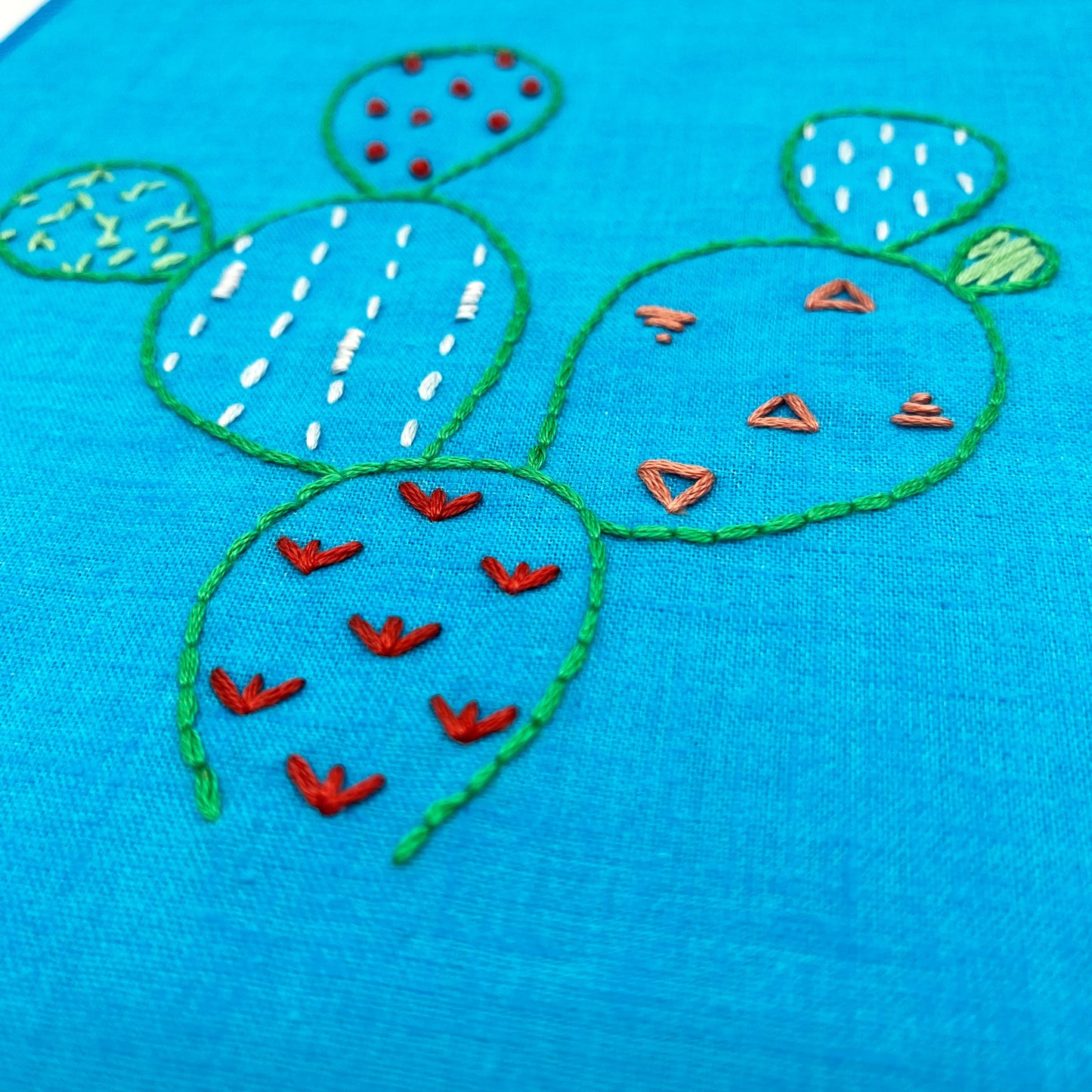 close up angled view of a bright blue fabric wall hanging, hand embroidered with the outline of a prickly pear cactus in green, each pad filled with different stitches like french knots, running stitches and triangles, in different colors