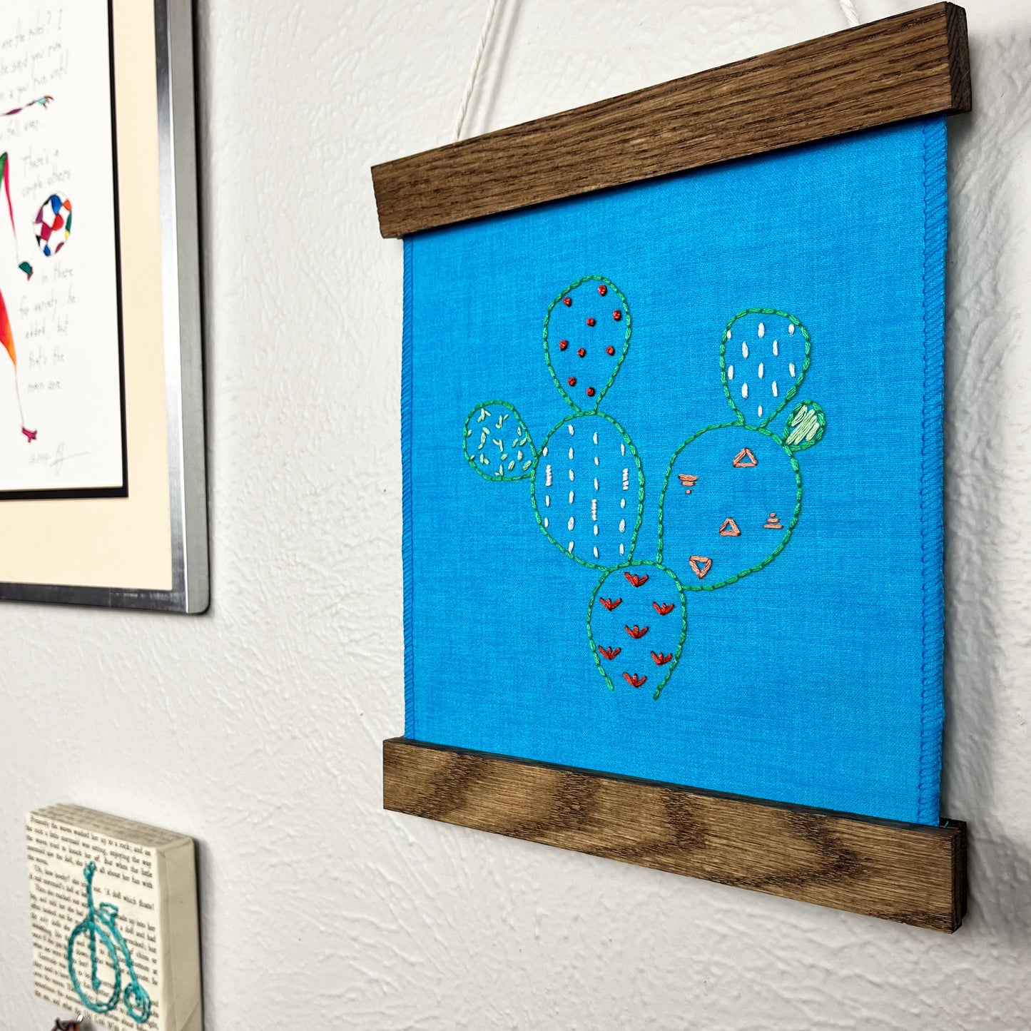 a small bright blue fabric wall hanging in a magnetic wood frame, hand embroidered with the outline of a prickly pear cactus in green, each pad filled with different stitches in different colors, hanging on a wall near other artwork