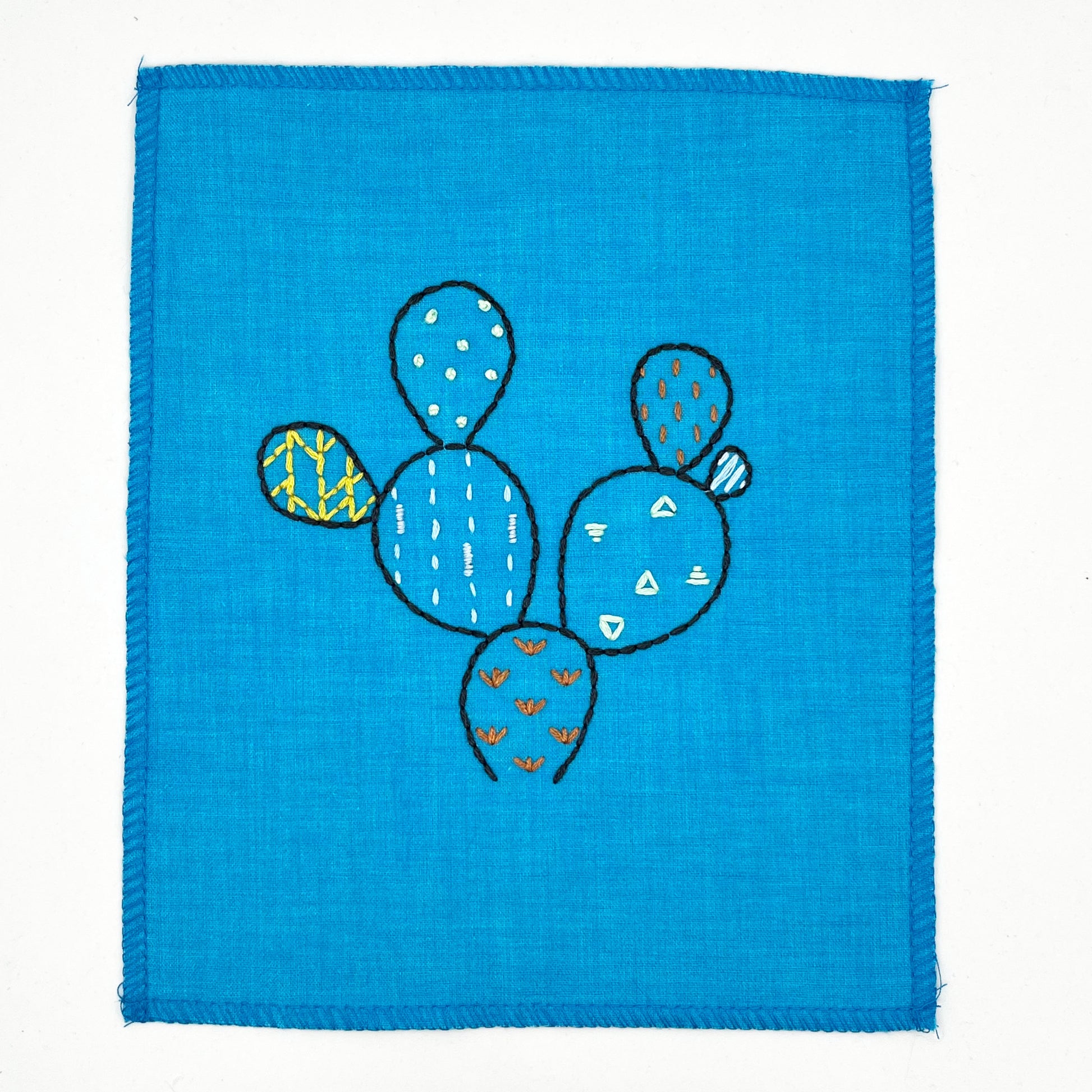 a small bright blue fabric wall hanging, hand embroidered with the outline of a prickly pear cactus in dark blue, each pad filled with different stitches like french knots, running stitches and triangles, in different colors, on a white background