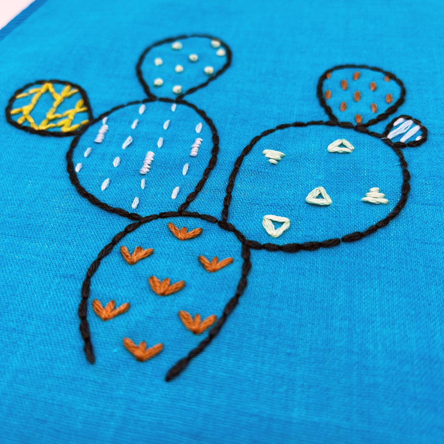 close up angled view of a bright blue fabric wall hanging, hand embroidered with the outline of a prickly pear cactus in dark blue, each pad filled with different stitches like french knots, running stitches and triangles, in different colors