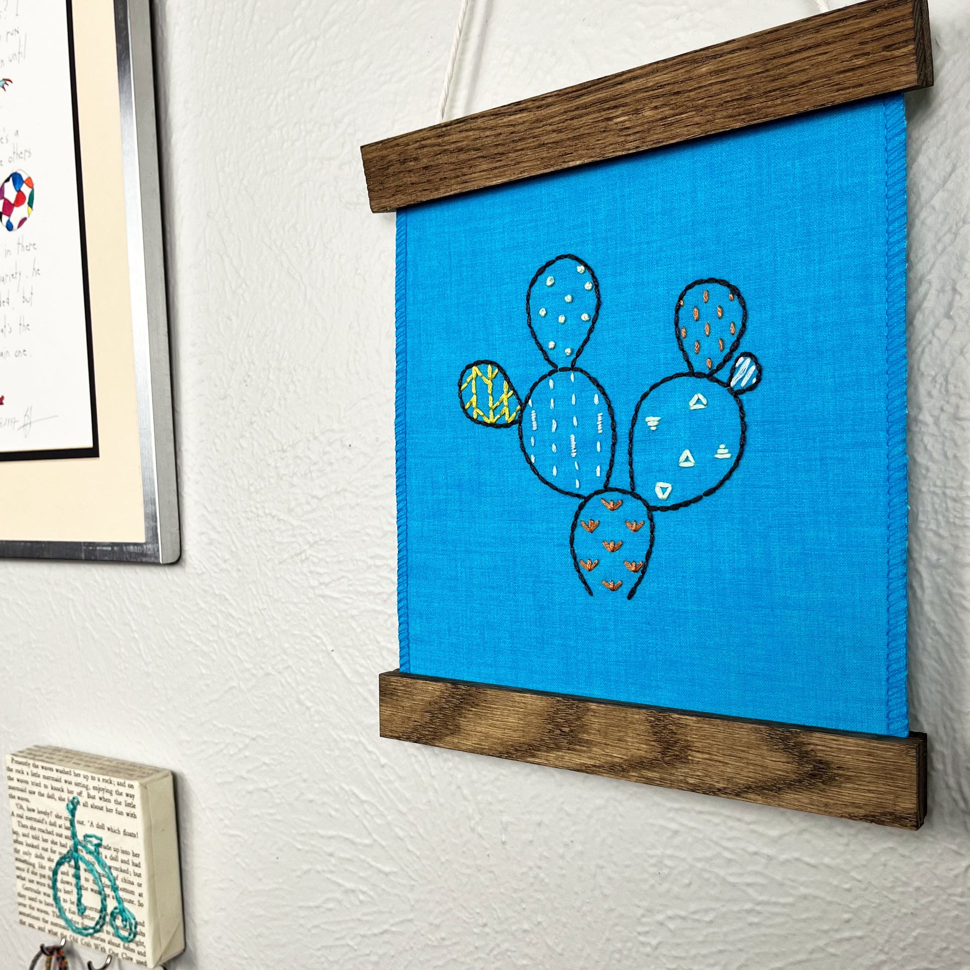 a small bright blue fabric wall hanging in a magnetic wood frame, hand embroidered with the outline of a prickly pear cactus in dark blue, each pad filled with different stitches in different colors, hanging on a wall near other artwork