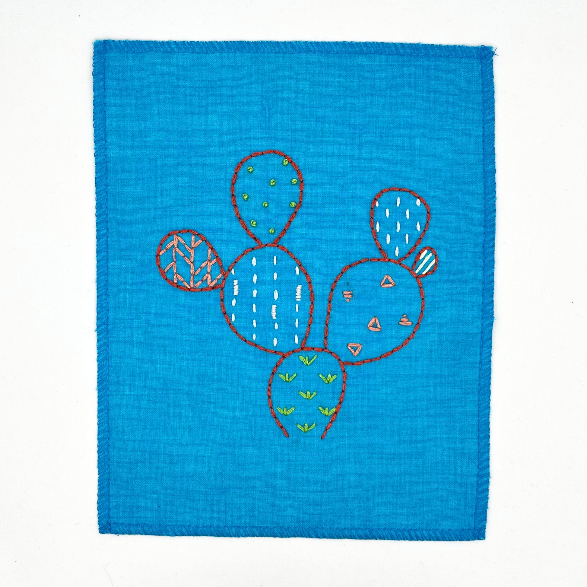 a small bright blue fabric wall hanging, hand embroidered with the outline of a prickly pear cactus in red, each pad filled with different stitches like french knots, running stitches and triangles, in different colors, on a white background