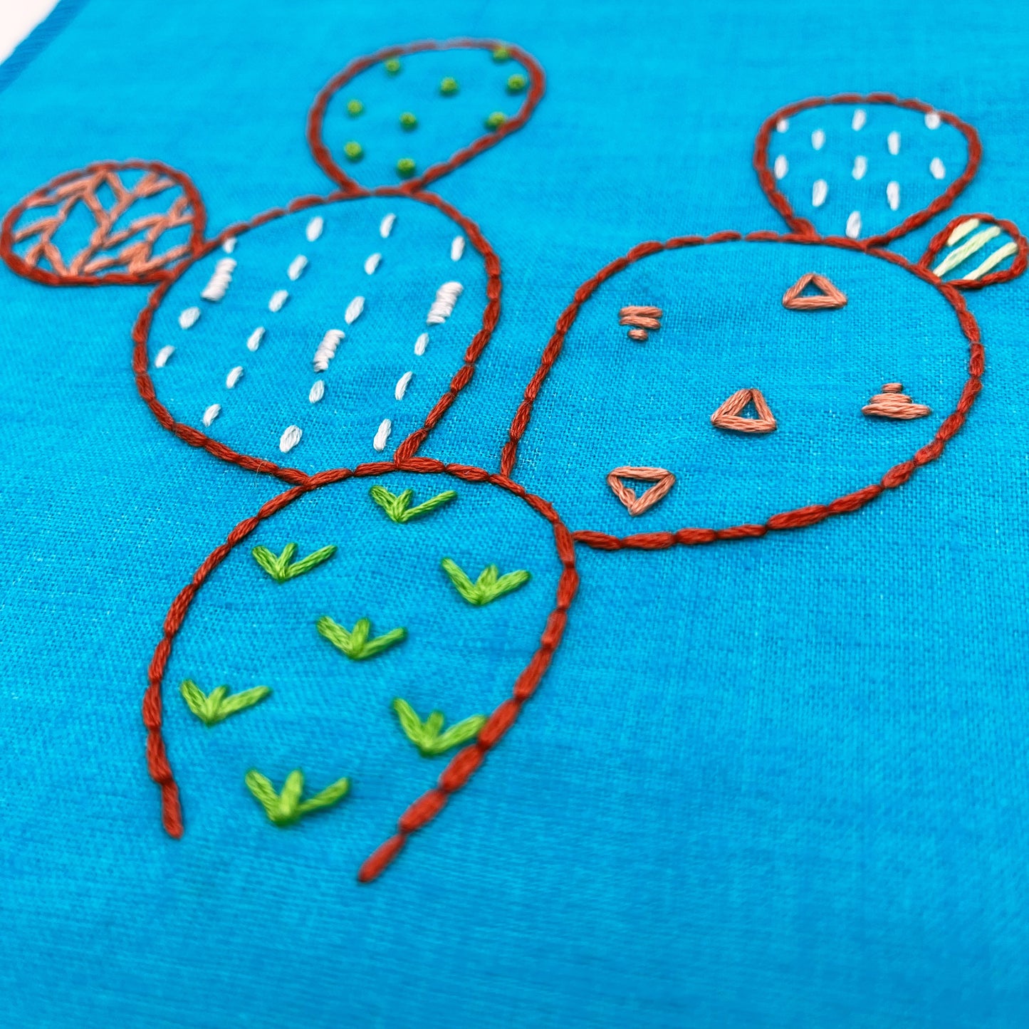 close up angled view of a bright blue fabric wall hanging, hand embroidered with the outline of a prickly pear cactus in red, each pad filled with different stitches like french knots, running stitches and triangles, in different colors