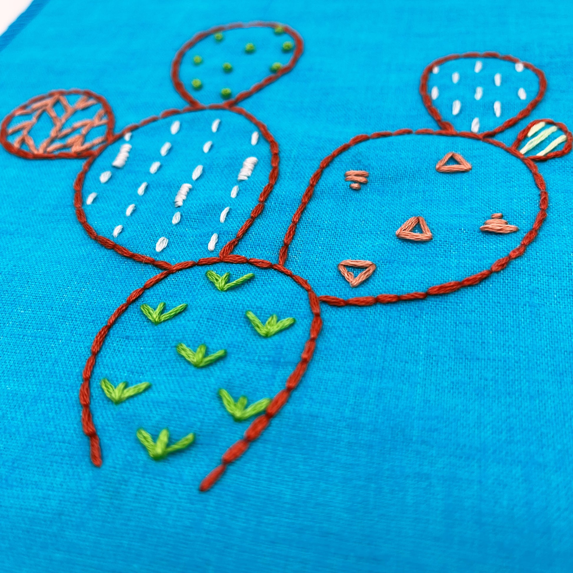 close up angled view of a bright blue fabric wall hanging, hand embroidered with the outline of a prickly pear cactus in red, each pad filled with different stitches like french knots, running stitches and triangles, in different colors