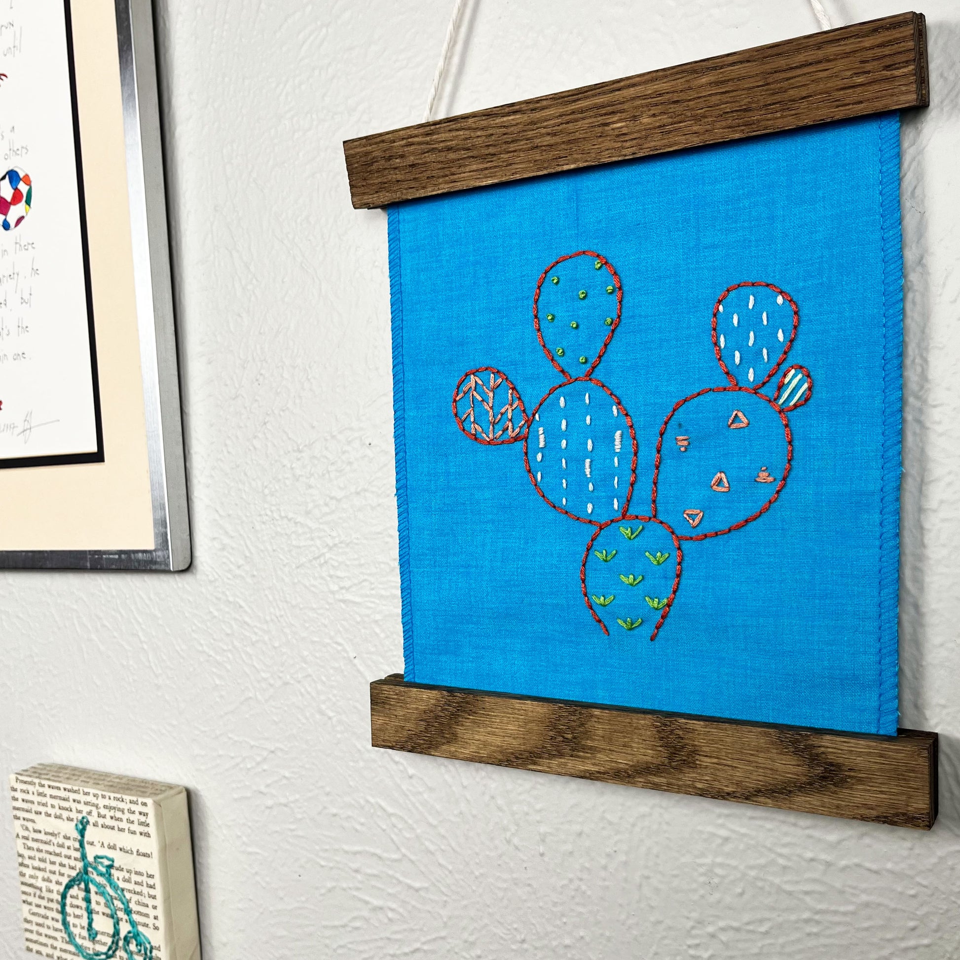 a small bright blue fabric wall hanging in a magnetic wood frame, hand embroidered with the outline of a prickly pear cactus in red, each pad filled with different stitches in different colors, hanging on a wall near other artwork