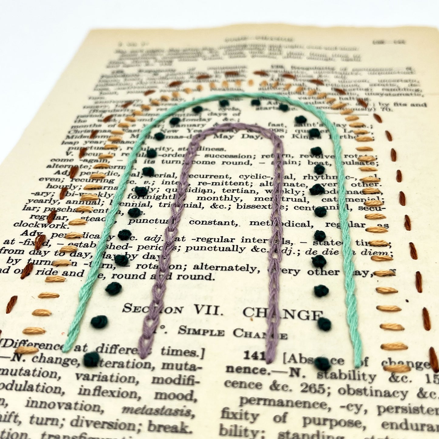 a close up angled view of a vintage book page, embroidered with a rainbow in different types of embroidery stitches, on a white background