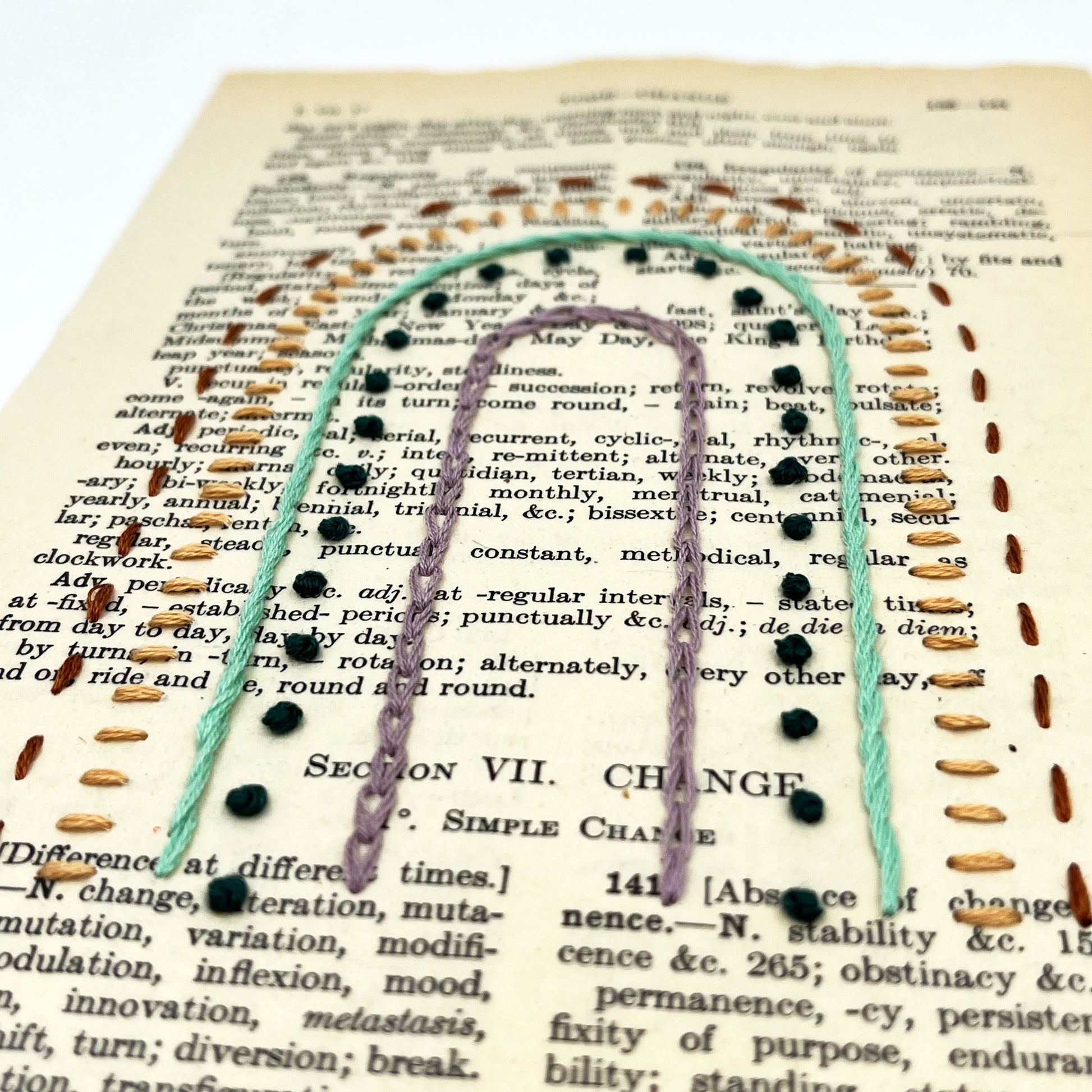 a close up angled view of a vintage book page, embroidered with a rainbow in different types of embroidery stitches, on a white background
