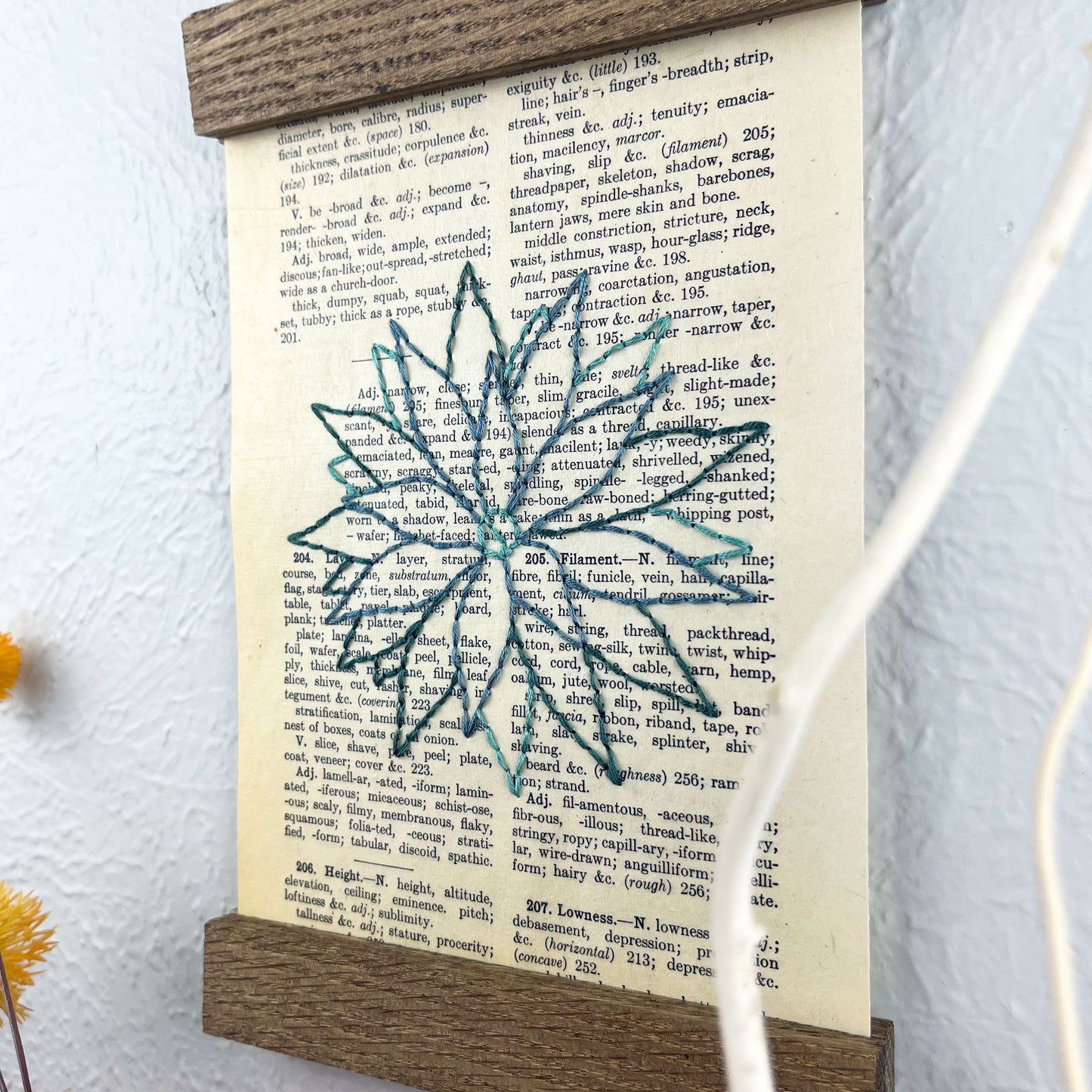 a vintage thesaurus page in a magnetic wood frame hanging on a wall, the book page is hand embroidered in green colored thread with an agave plant