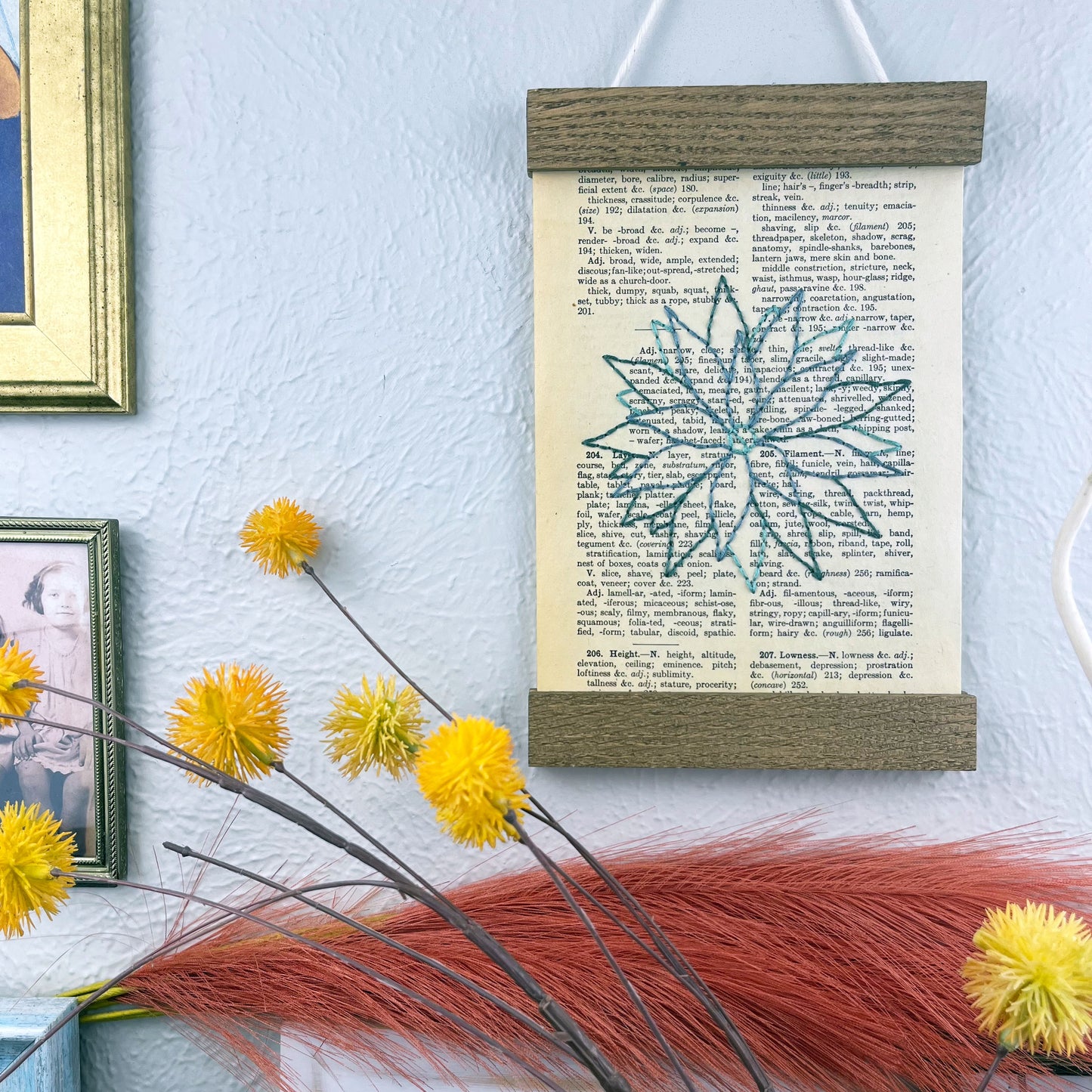 a vintage thesaurus page in a magnetic wood frame hanging on a wall next to framed photos, decorative branches and feathers, the book page is hand embroidered in green colored thread with an agave plant