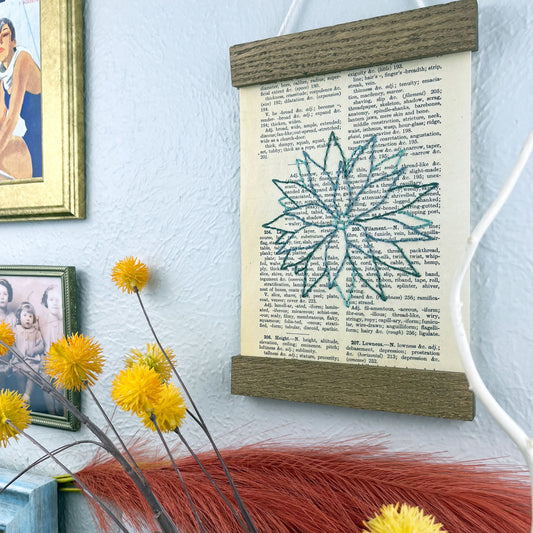 a vintage thesaurus page in a magnetic wood frame hanging on a wall next to framed photos, decorative branches and feathers, the book page is hand embroidered in green colored thread with an agave plant