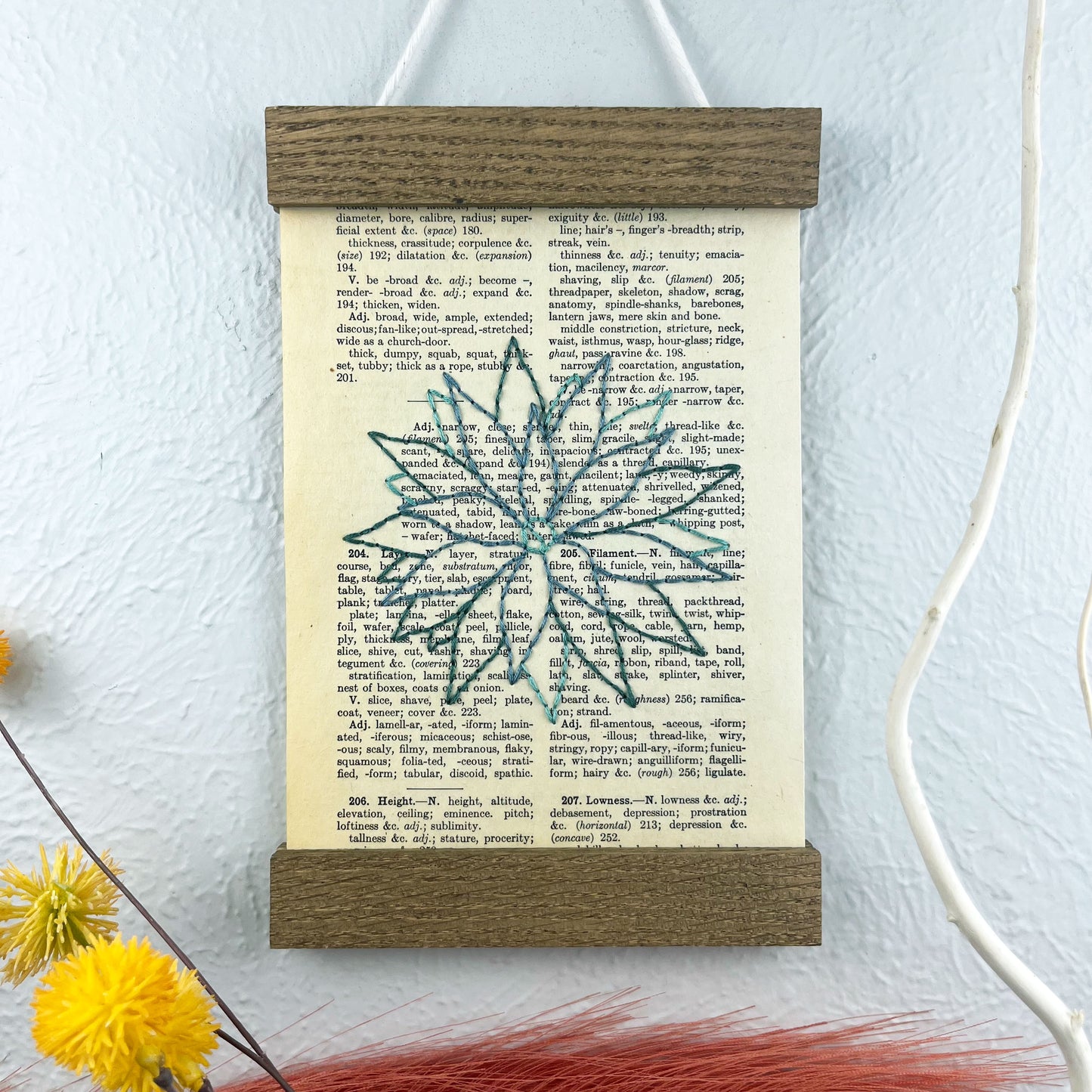 a vintage thesaurus page in a magnetic wood frame hanging on a wall next to  decorative branches and feathers, the book page is hand embroidered in green colored thread with an agave plant