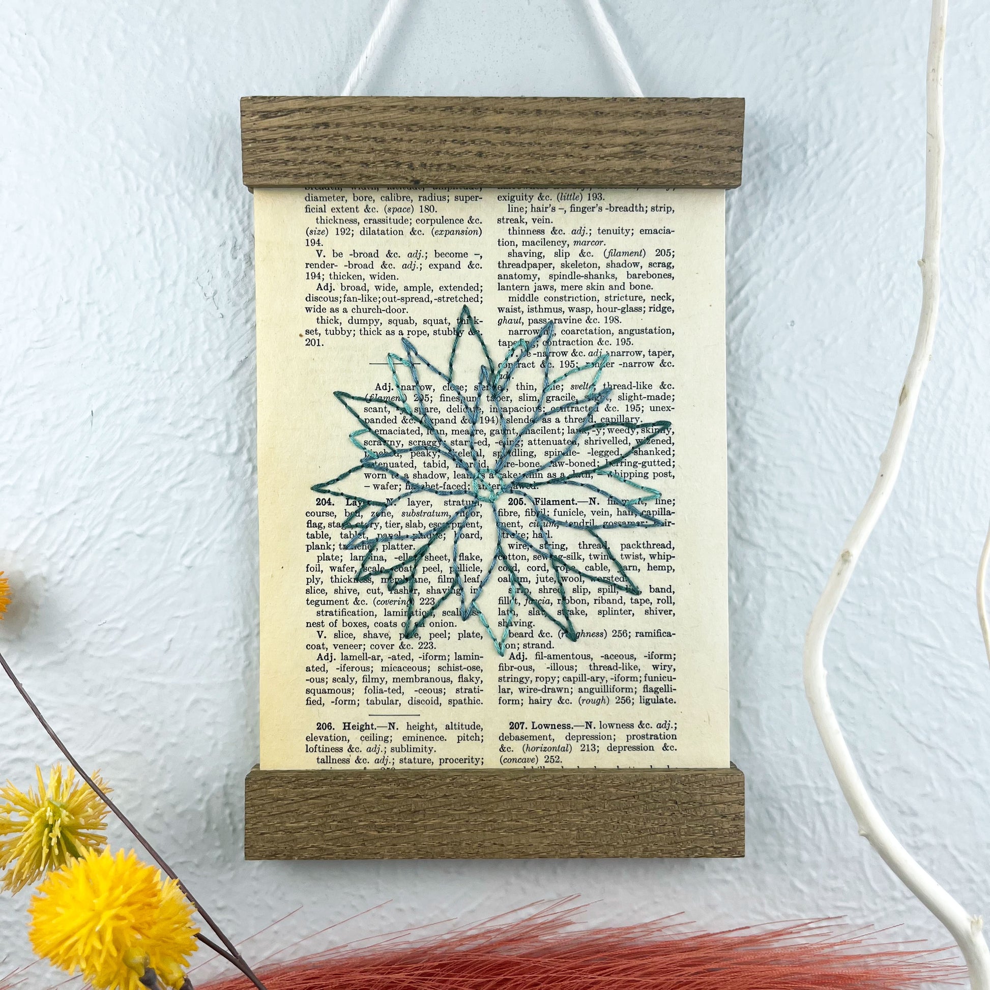 a vintage thesaurus page in a magnetic wood frame hanging on a wall next to  decorative branches and feathers, the book page is hand embroidered in green colored thread with an agave plant