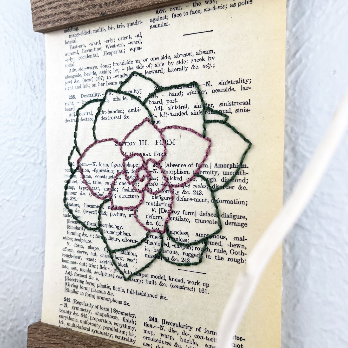 a vintage thesaurus page in a magnetic wood frame hanging on a wall, the book page is hand embroidered in purply pink and green colored thread with an echeveria succulent