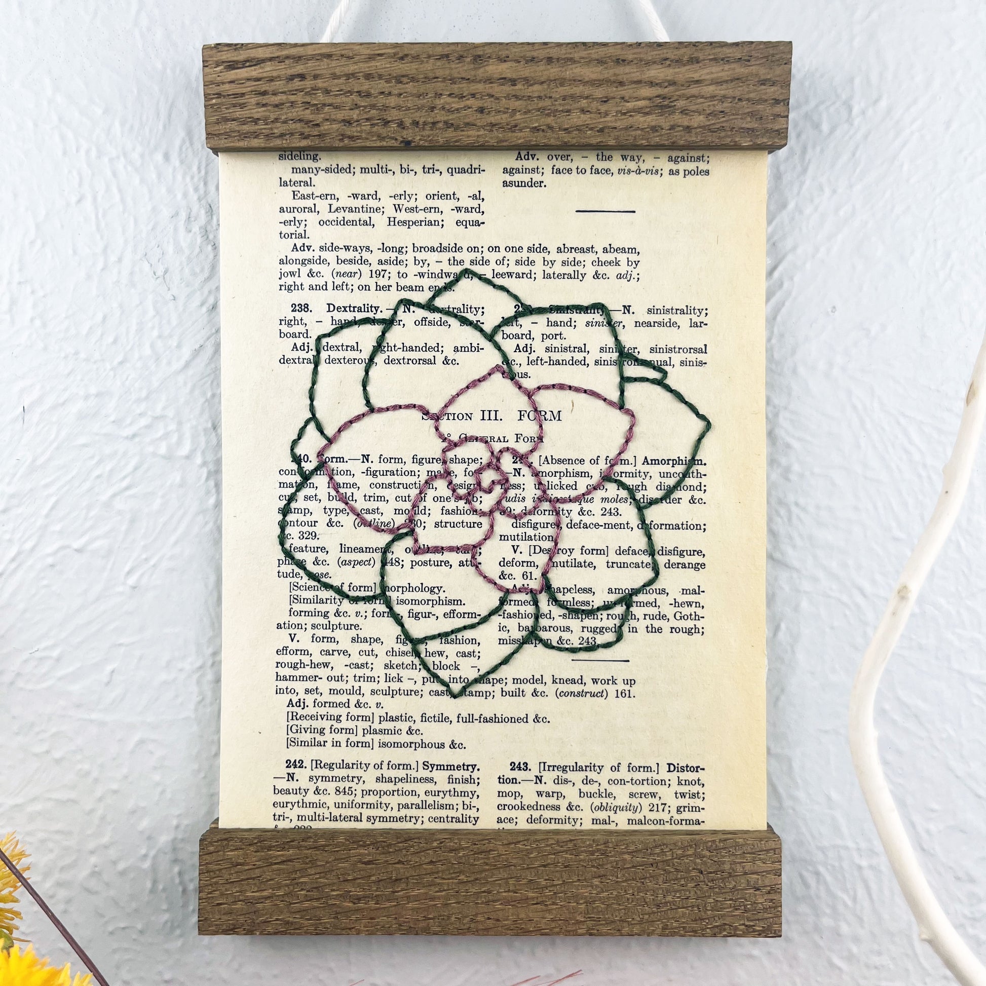 a vintage thesaurus page in a magnetic wood frame hanging on a wall, the book page is hand embroidered in purply pink and green colored thread with an echeveria succulent