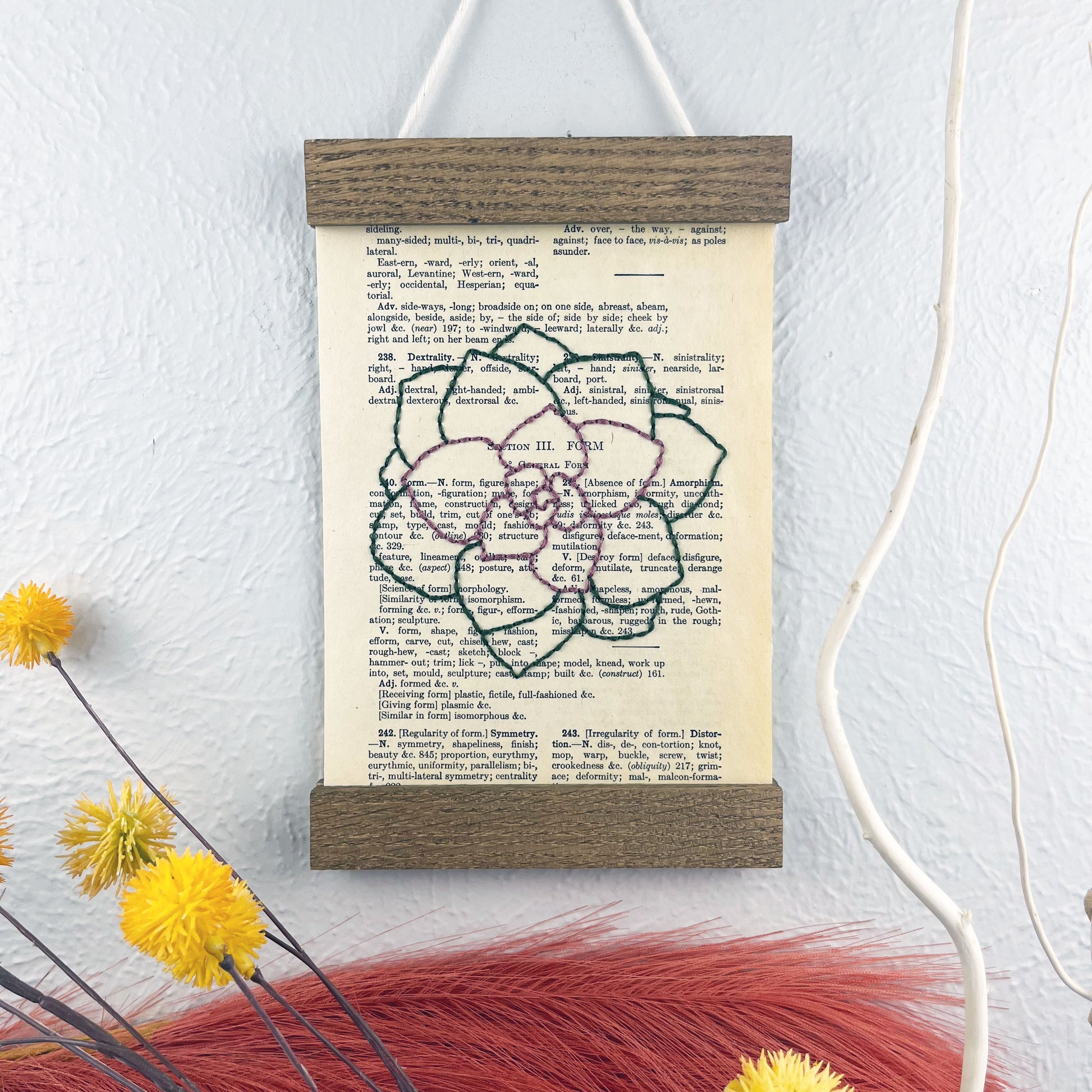 a vintage thesaurus page in a magnetic wood frame hanging on a wall next to framed photos, decorative branches and feathers, the book page is hand embroidered in purply pink and green colored thread with an echeveria succulent