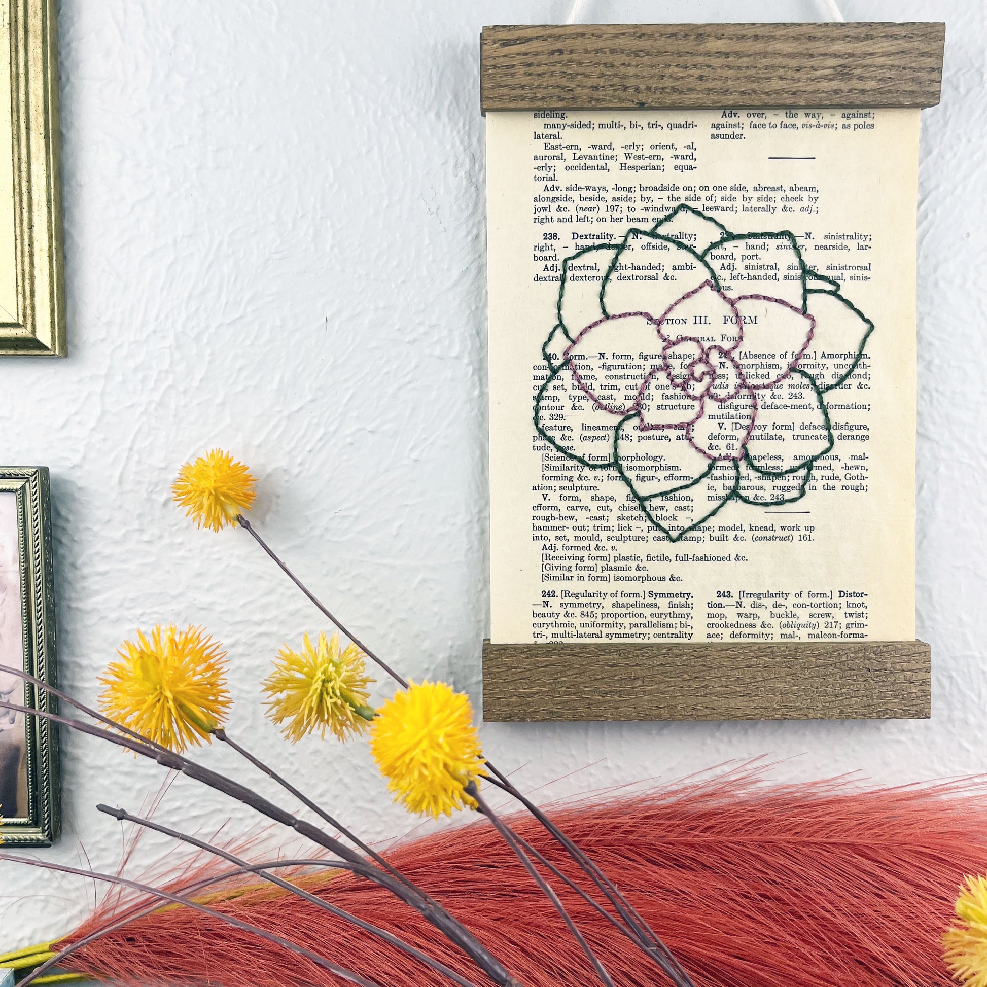 a vintage thesaurus page in a magnetic wood frame hanging on a wall next to framed photos, decorative branches and feathers, the book page is hand embroidered in purply pink and green colored thread with an echeveria succulent
