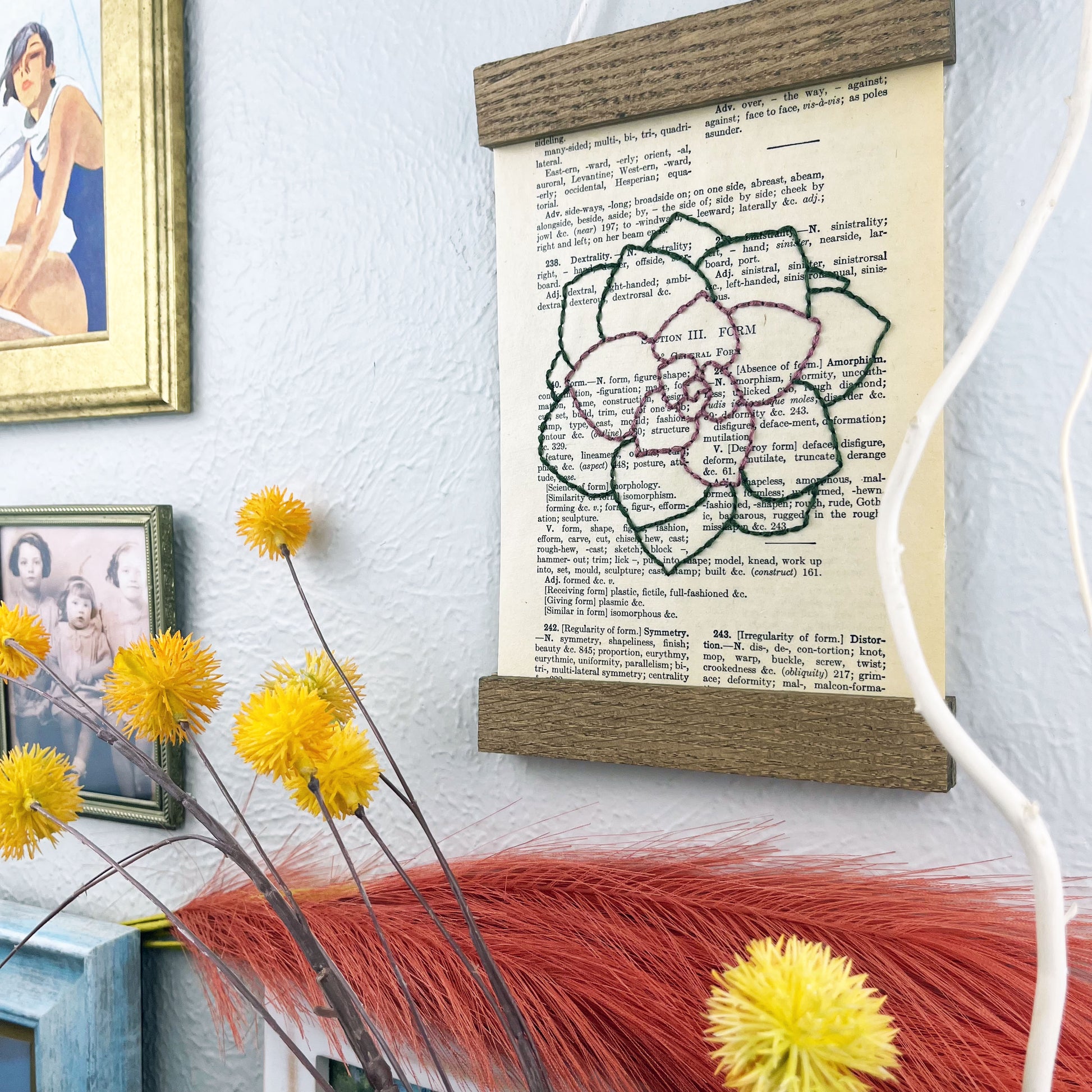 a vintage thesaurus page in a magnetic wood frame hanging on a wall next to framed photos, decorative branches and feathers, the book page is hand embroidered in purply pink and green colored thread with an echeveria succulent