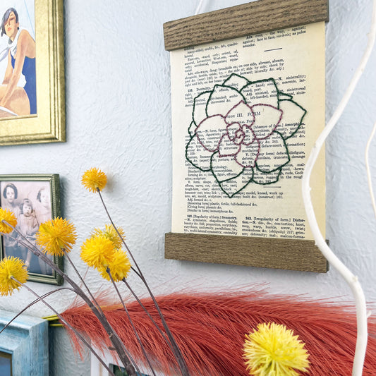 a vintage thesaurus page in a magnetic wood frame hanging on a wall next to framed photos, decorative branches and feathers, the book page is hand embroidered in purply pink and green colored thread with an echeveria succulent