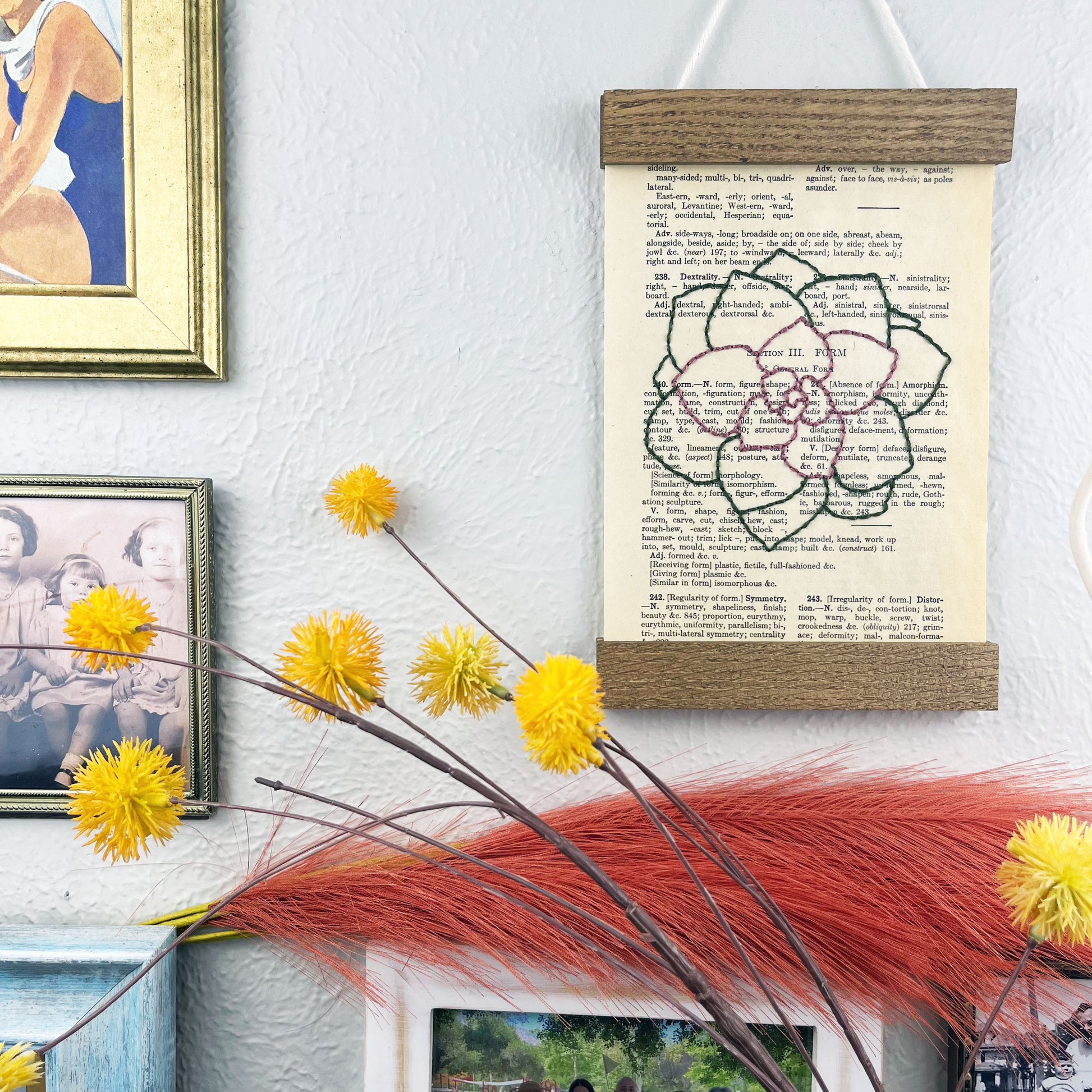 a vintage thesaurus page in a magnetic wood frame hanging on a wall next to framed photos, decorative branches and feathers, the book page is hand embroidered in purply pink and green colored thread with an echeveria succulent