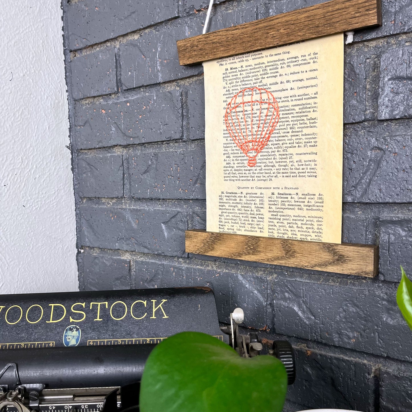 a vintage thesaurus page, hand stitched over with an outline of a hot air balloon in coral thread, hanging in a magnetic wood frame on a grey brick wall, above a typewriter and a pothos plant