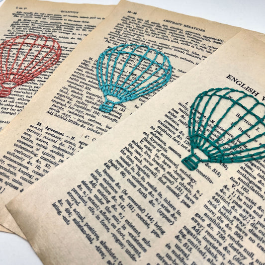 close up angled view of three loose pages from a vintage thesaurus, with three different colored outlines of hot air balloons stitched through them- coral, robin's egg blue and teal