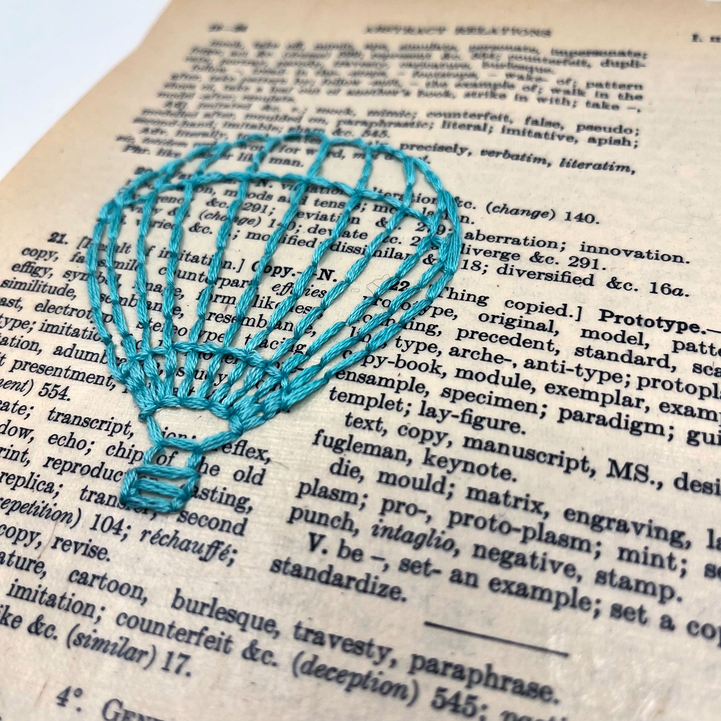 a close up angled view of a vintage thesaurus page, hand stitched over with an outline of a hot air balloon in robin's egg blue thread