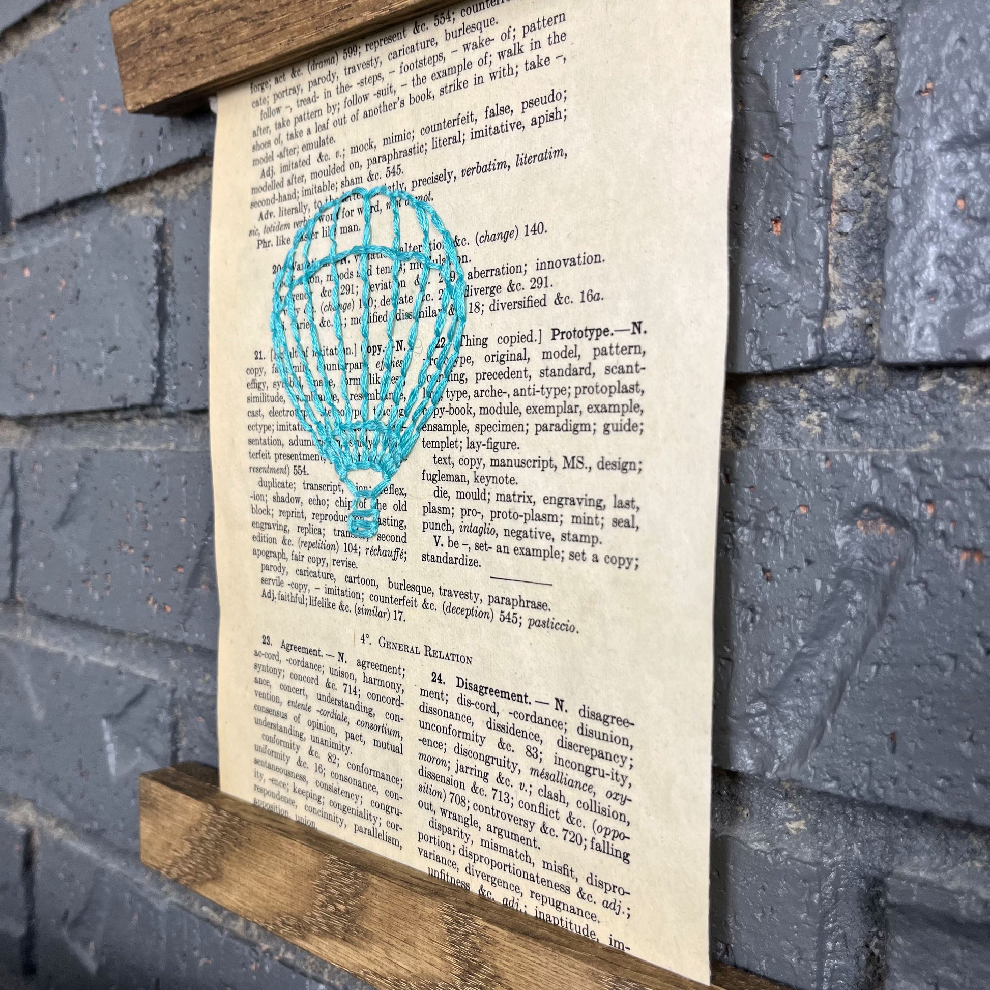 a close up angled view of a vintage thesaurus page, hand stitched over with an outline of a hot air balloon in robin's egg blue thread, hanging in a magnetic wood frame on a grey brick wall