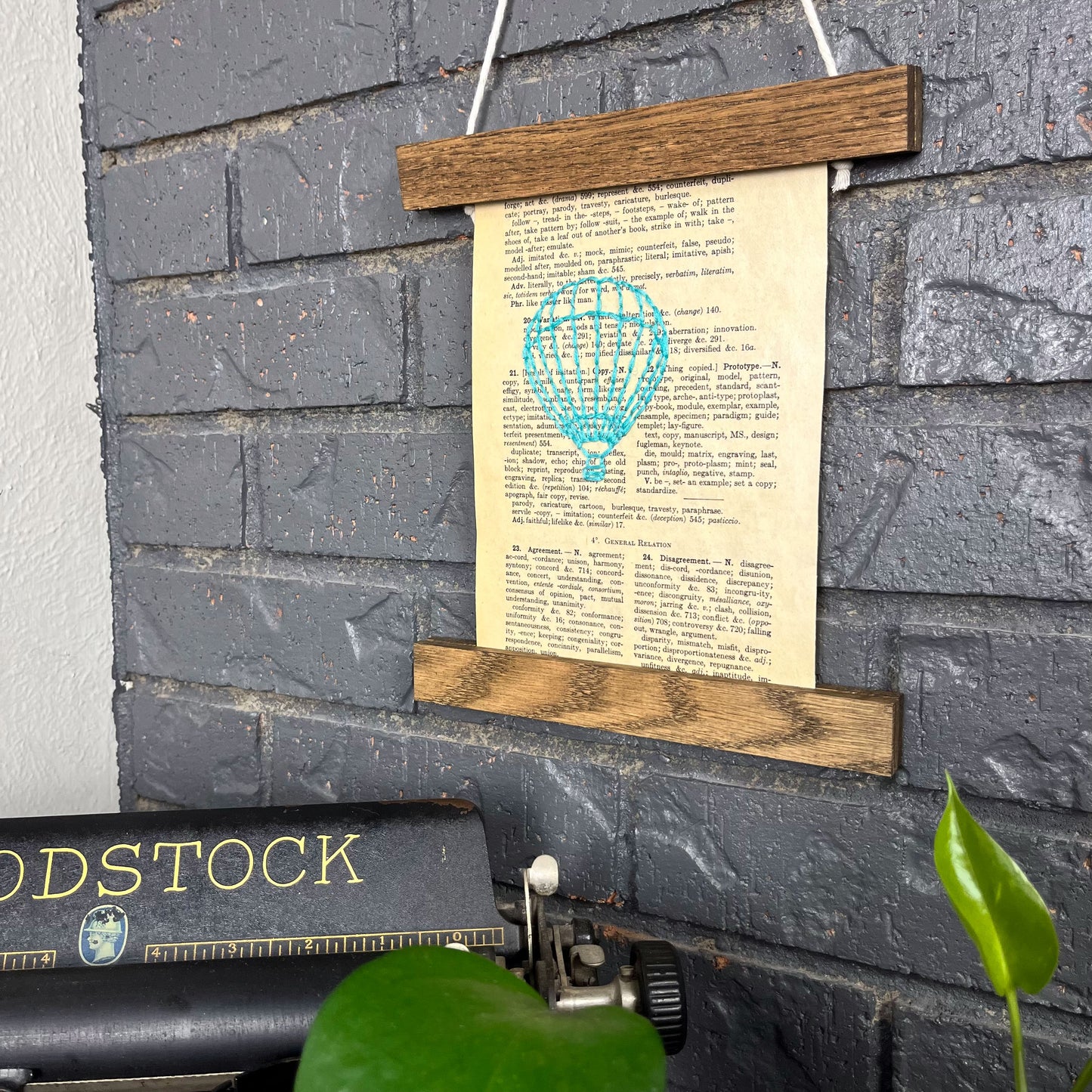 a vintage thesaurus page, hand stitched over with an outline of a hot air balloon in robin's egg blue thread, hanging in a magnetic wood frame on a grey brick wall, above a typewriter and a pothos plant