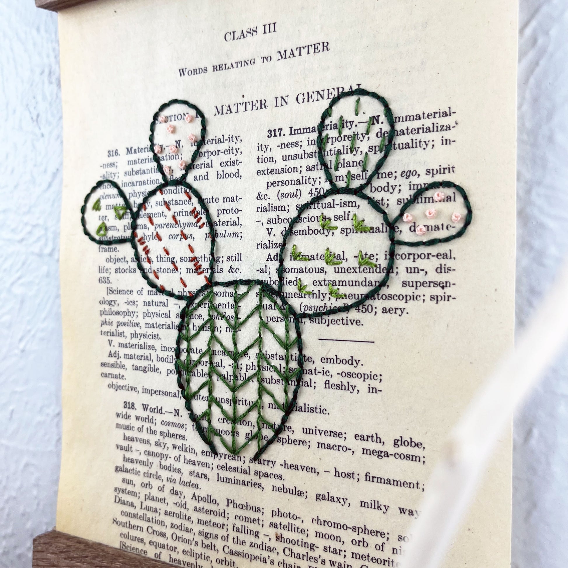 close up view of a vintage thesaurus page in a magnetic wood frame hanging on a wall, the book page is hand embroidered in green peach and rust colored thread with a prickly pear cactus with different patterns filled in each pad