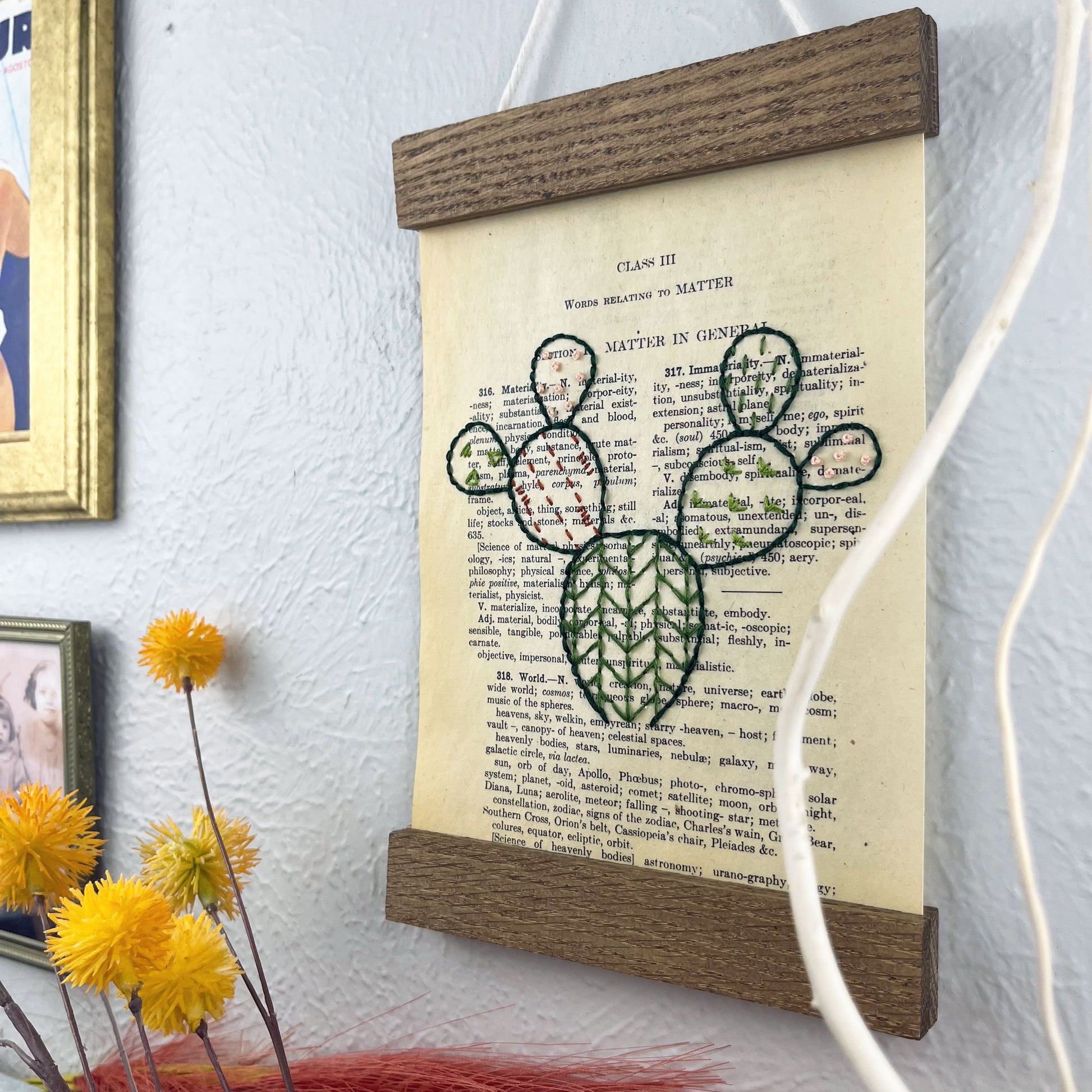 a vintage thesaurus page in a magnetic wood frame hanging on a wall next to framed photos, decorative branches and feathers, the book page is hand embroidered in green peach and rust colored thread with a prickly pear cactus with different patterns filled in each pad