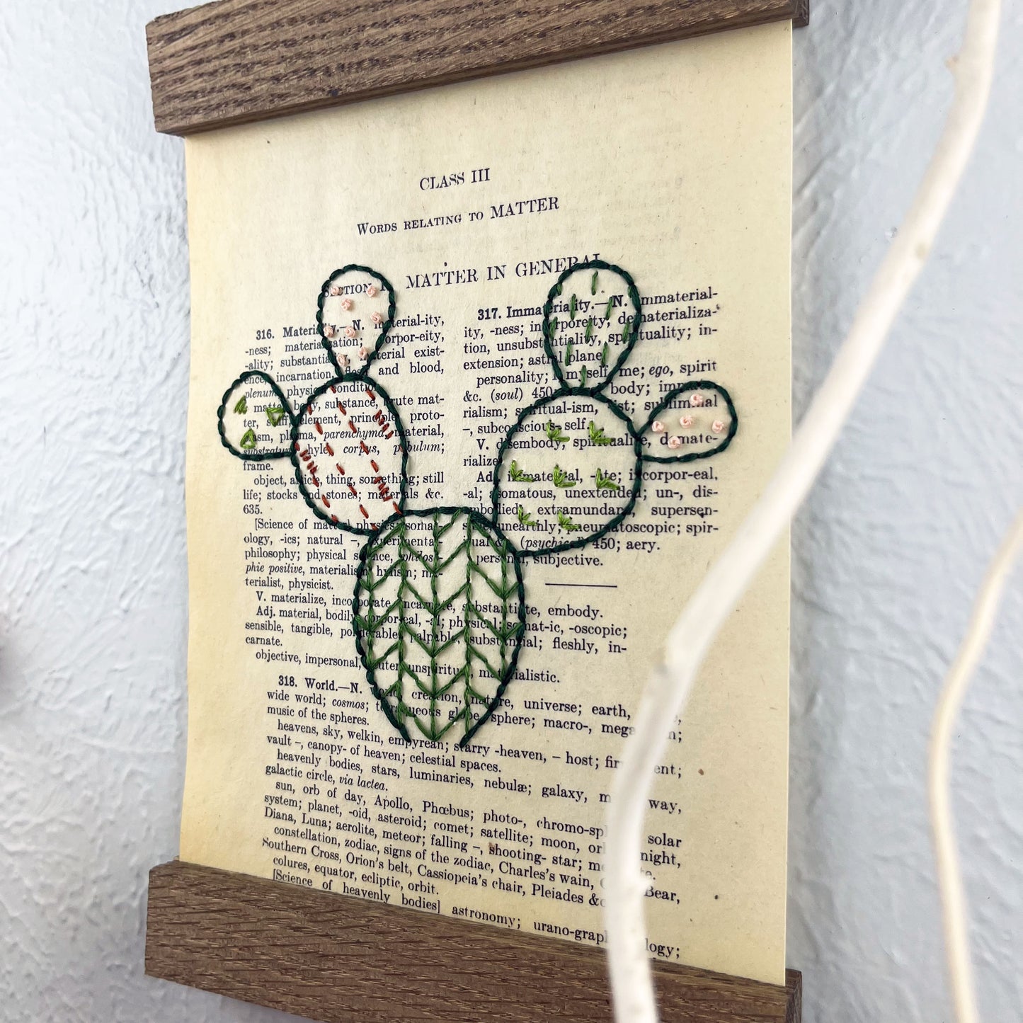 close up view of a vintage thesaurus page in a magnetic wood frame hanging on a wall, the book page is hand embroidered in green peach and rust colored thread with a prickly pear cactus with different patterns filled in each pad