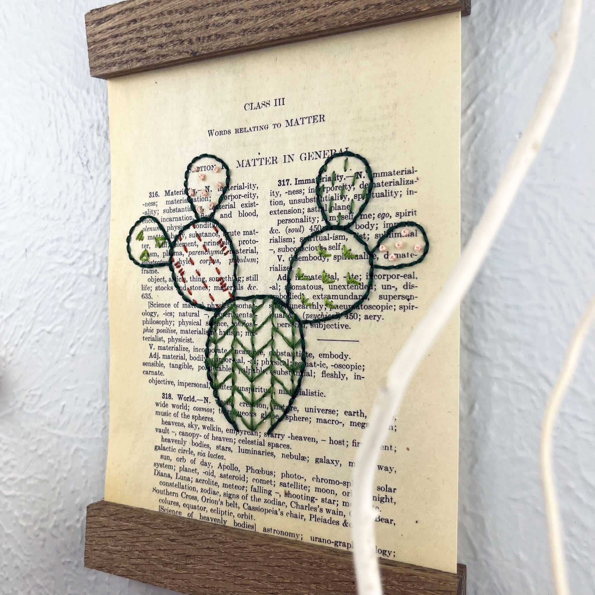 close up view of a vintage thesaurus page in a magnetic wood frame hanging on a wall, the book page is hand embroidered in green peach and rust colored thread with a prickly pear cactus with different patterns filled in each pad