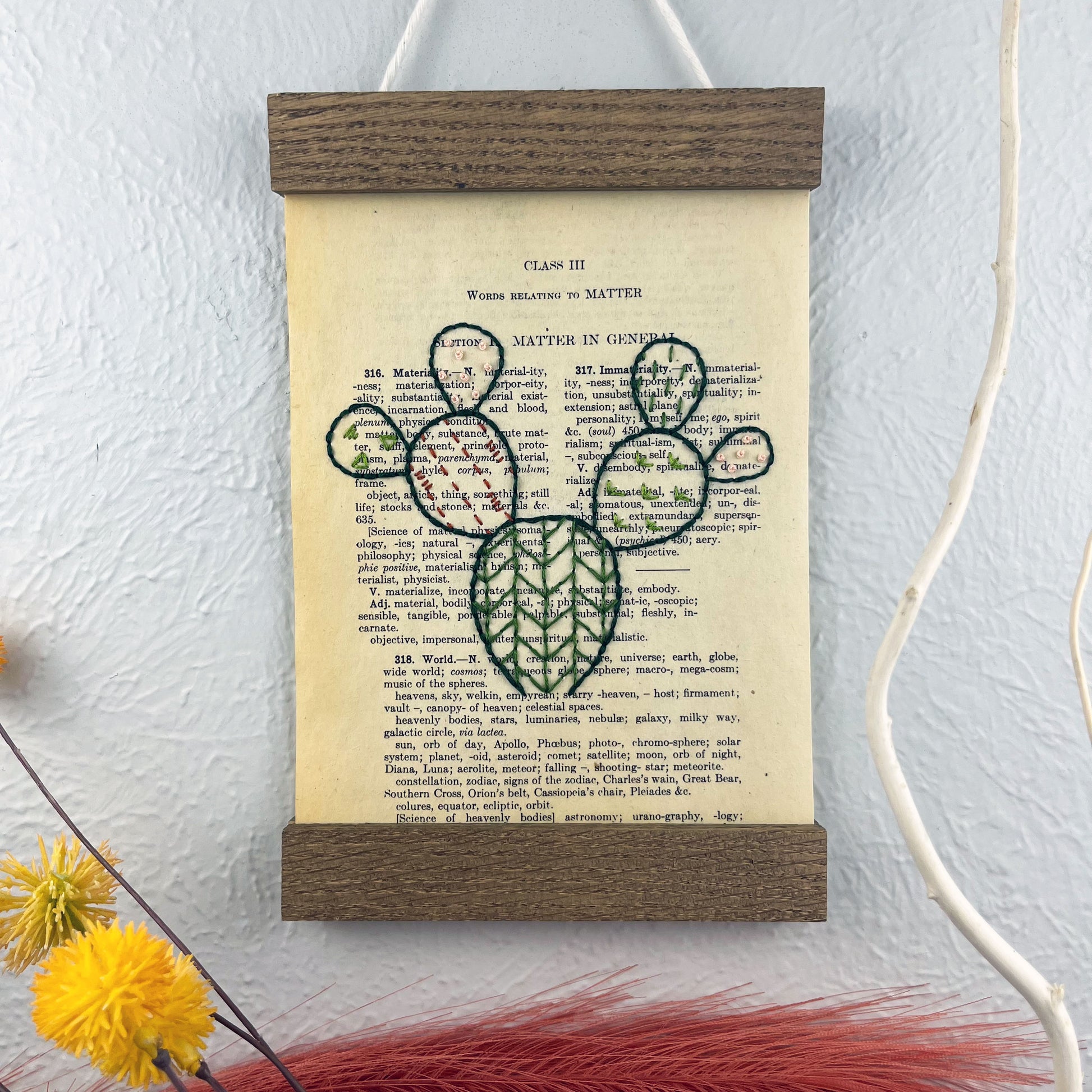 a vintage thesaurus page in a magnetic wood frame hanging on a wall next to  decorative branches and feathers, the book page is hand embroidered in green peach and rust colored thread with a prickly pear cactus with different patterns filled in each pad
