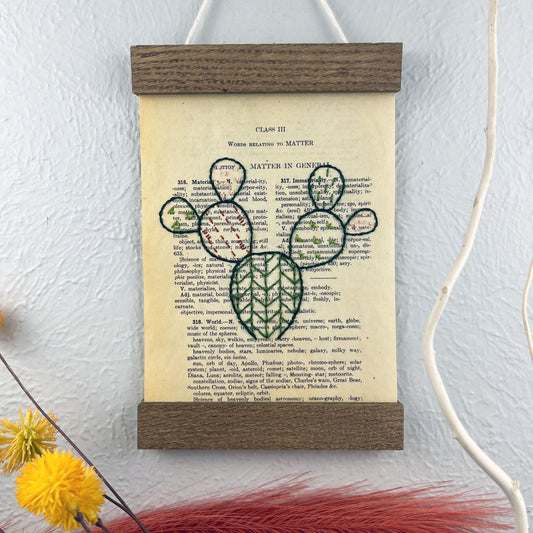 a vintage thesaurus page in a magnetic wood frame hanging on a wall next to  decorative branches and feathers, the book page is hand embroidered in green peach and rust colored thread with a prickly pear cactus with different patterns filled in each pad