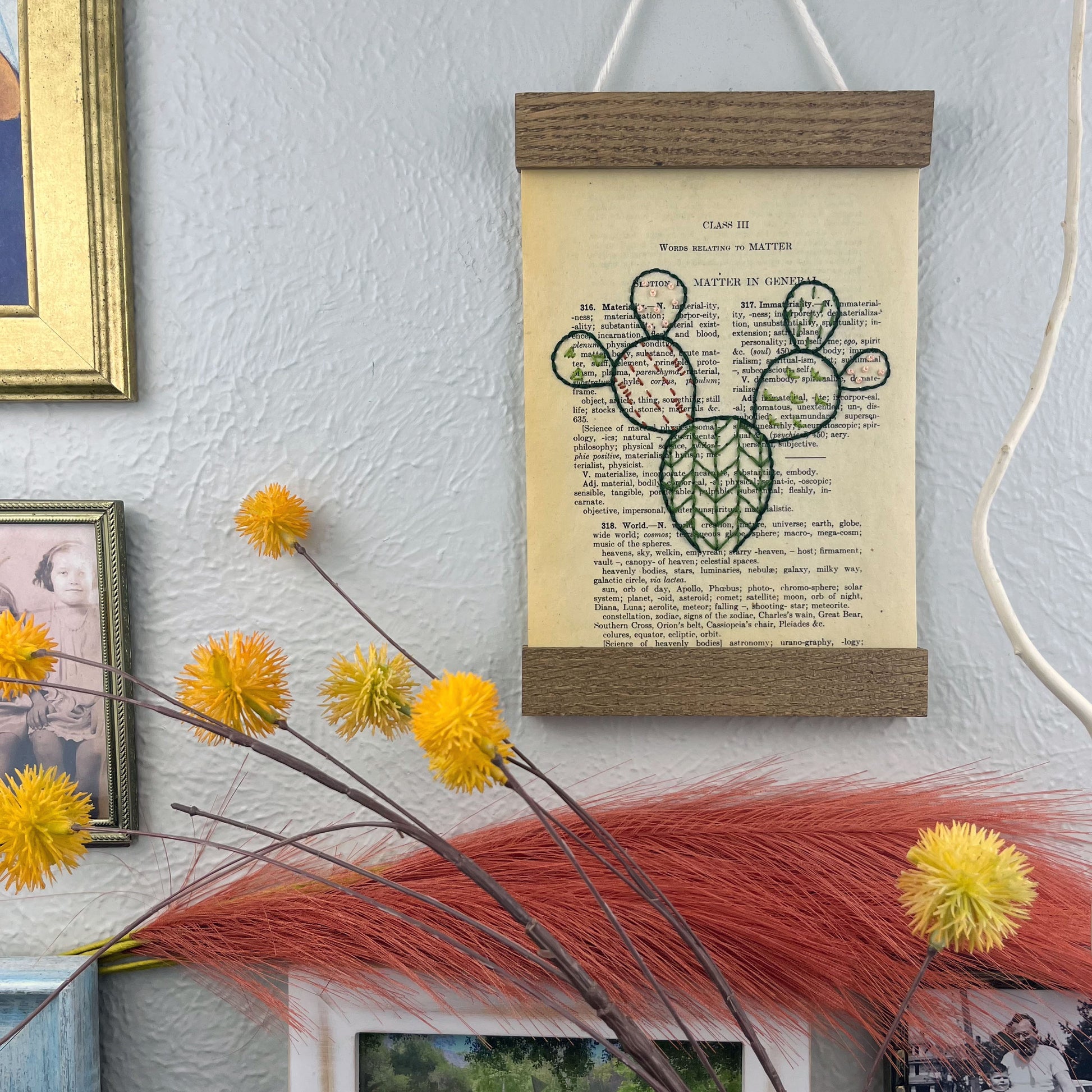 a vintage thesaurus page in a magnetic wood frame hanging on a wall next to framed photos, decorative branches and feathers, the book page is hand embroidered in green peach and rust colored thread with a prickly pear cactus with different patterns filled in each pad