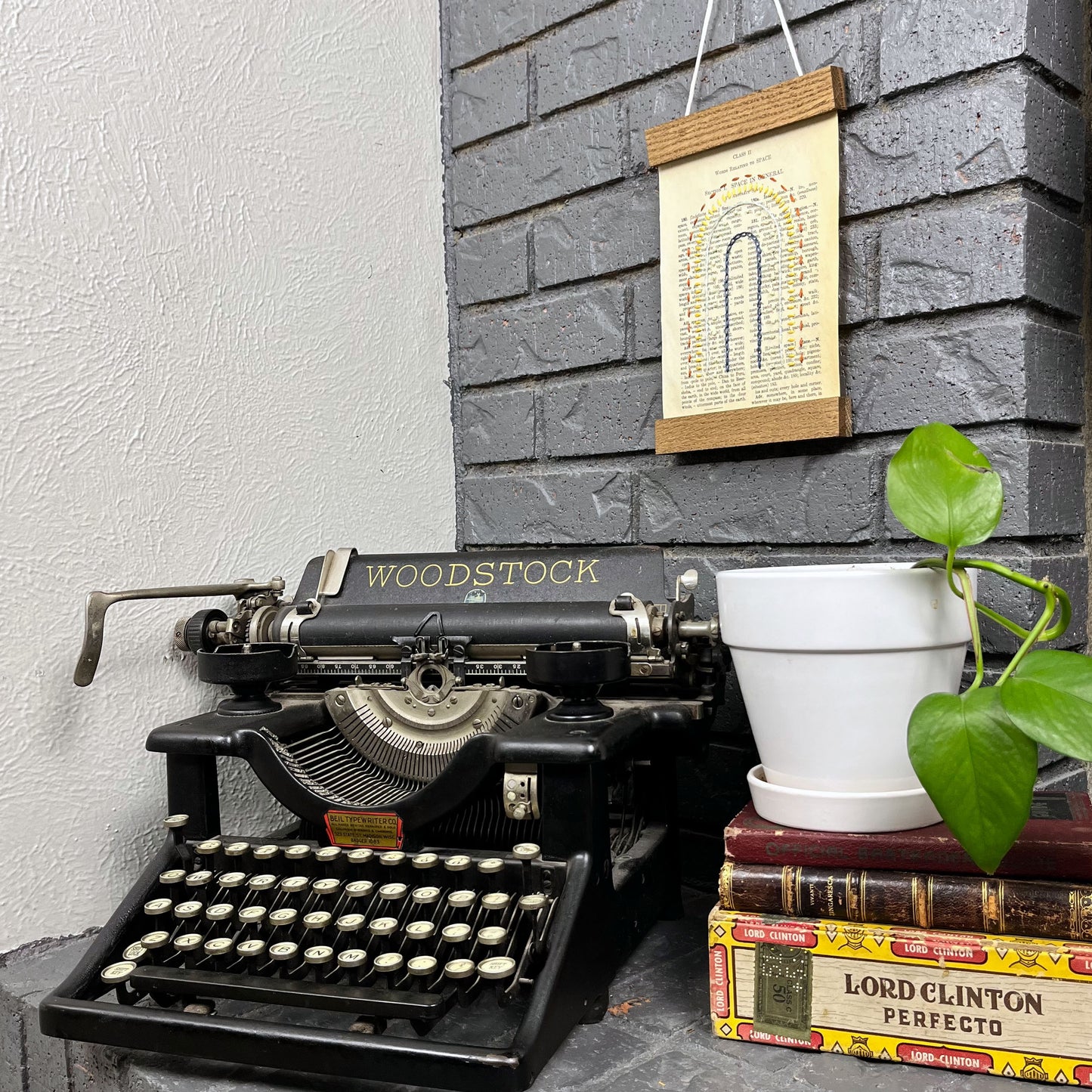 Vintage thesaurus page, embroidered with a rainbow in different types of stitches. Hanging in a magnetic wood frame on a grey brick wall, with a typewriter and pothos plant at the bottom of the picture