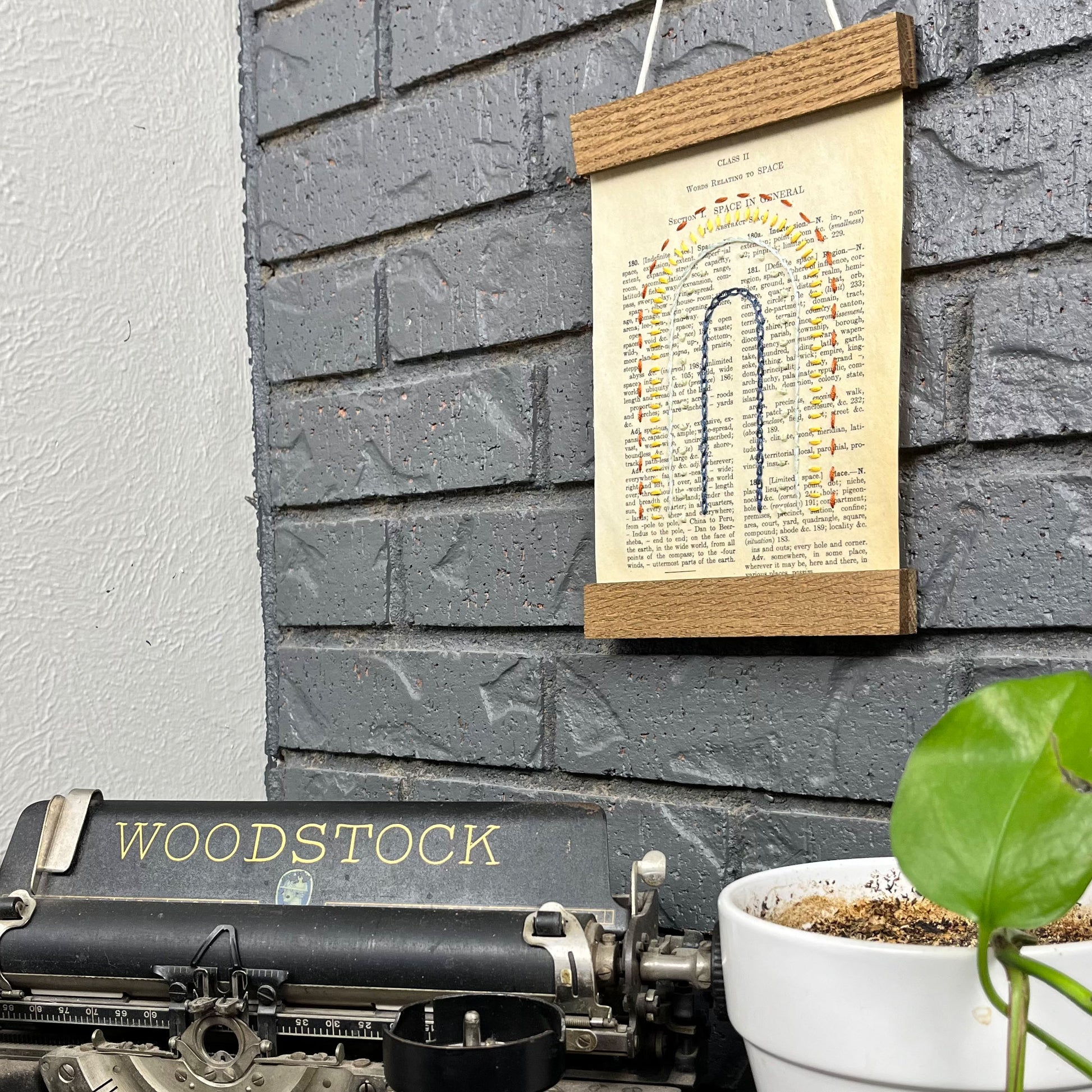 Vintage thesaurus page, embroidered with a rainbow in different types of stitches. Hanging in a magnetic wood frame on a grey brick wall, with the top of a typewriter and pothos plant peeking out at the bottom of the picture